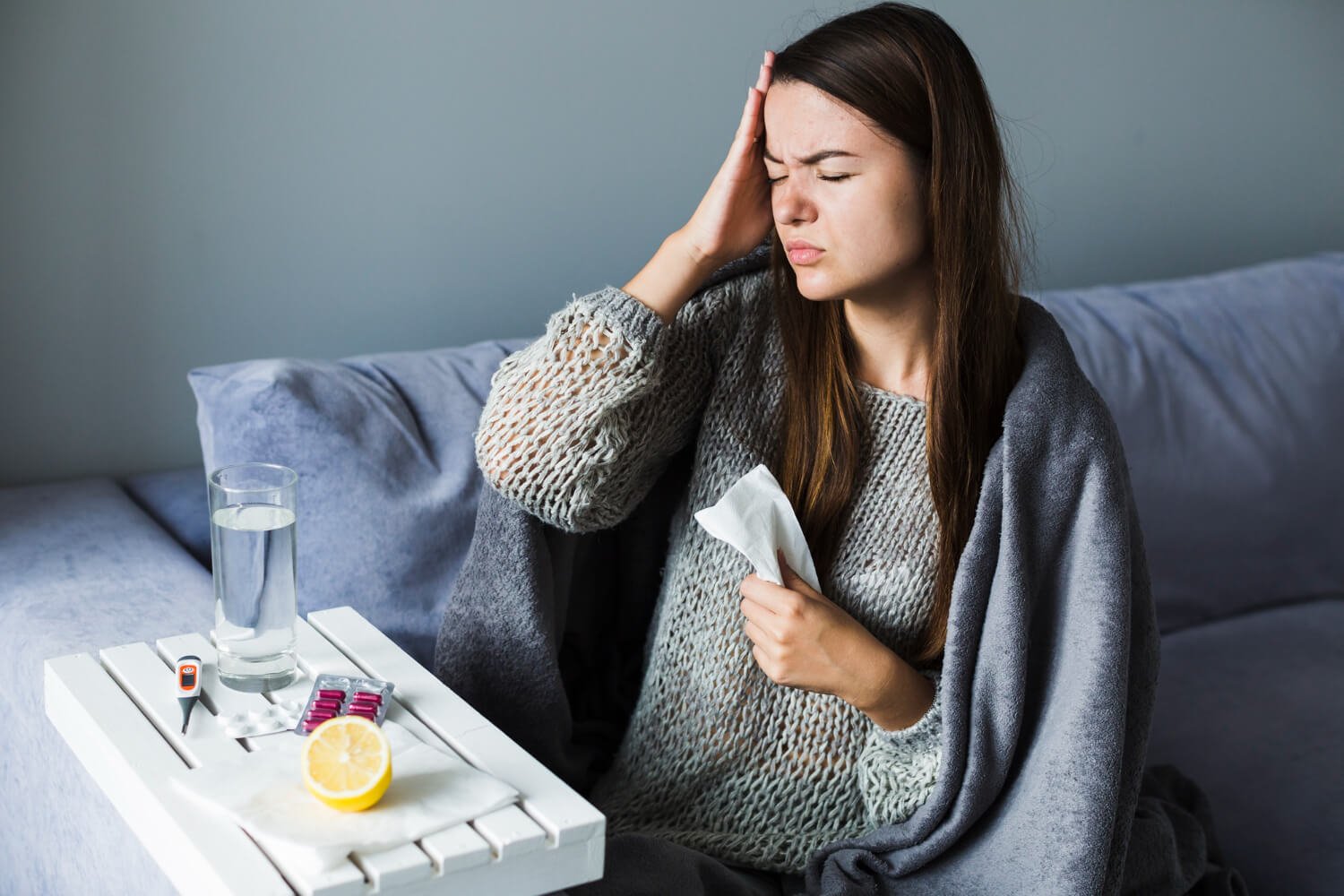 A woman sitting on a bed with a gray blanket, holding a tissue to her nose with a pained expression, indicating illness. On a white tray in front of her are a glass of water, a digital thermometer, a blister pack of pink pills, and half a lemon.