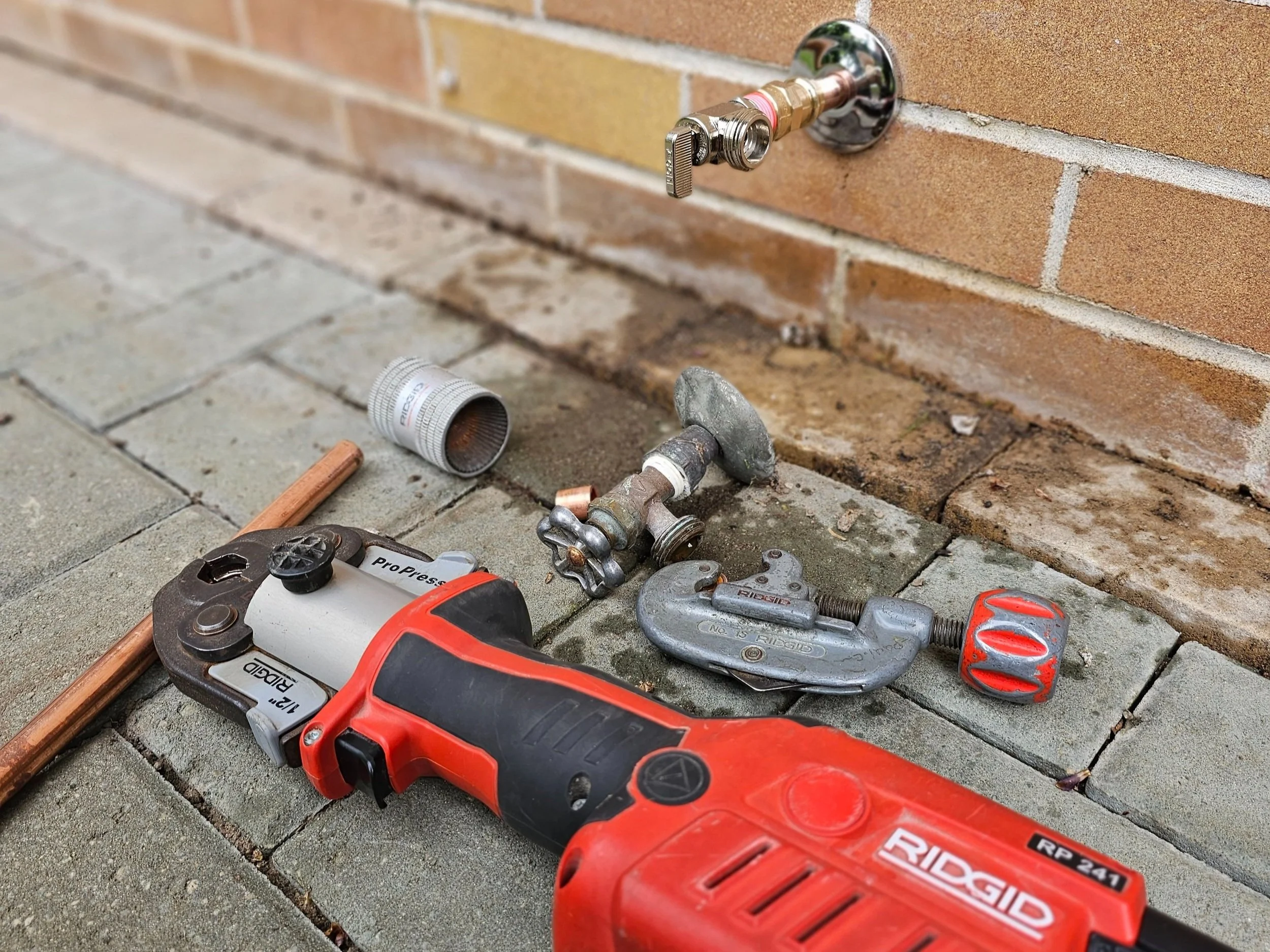 Tools and supplies for plumbing work on a brick wall outside, including a cordless ratchet wrench, a pipe clamp, a small hammer, and various pipes and fittings.