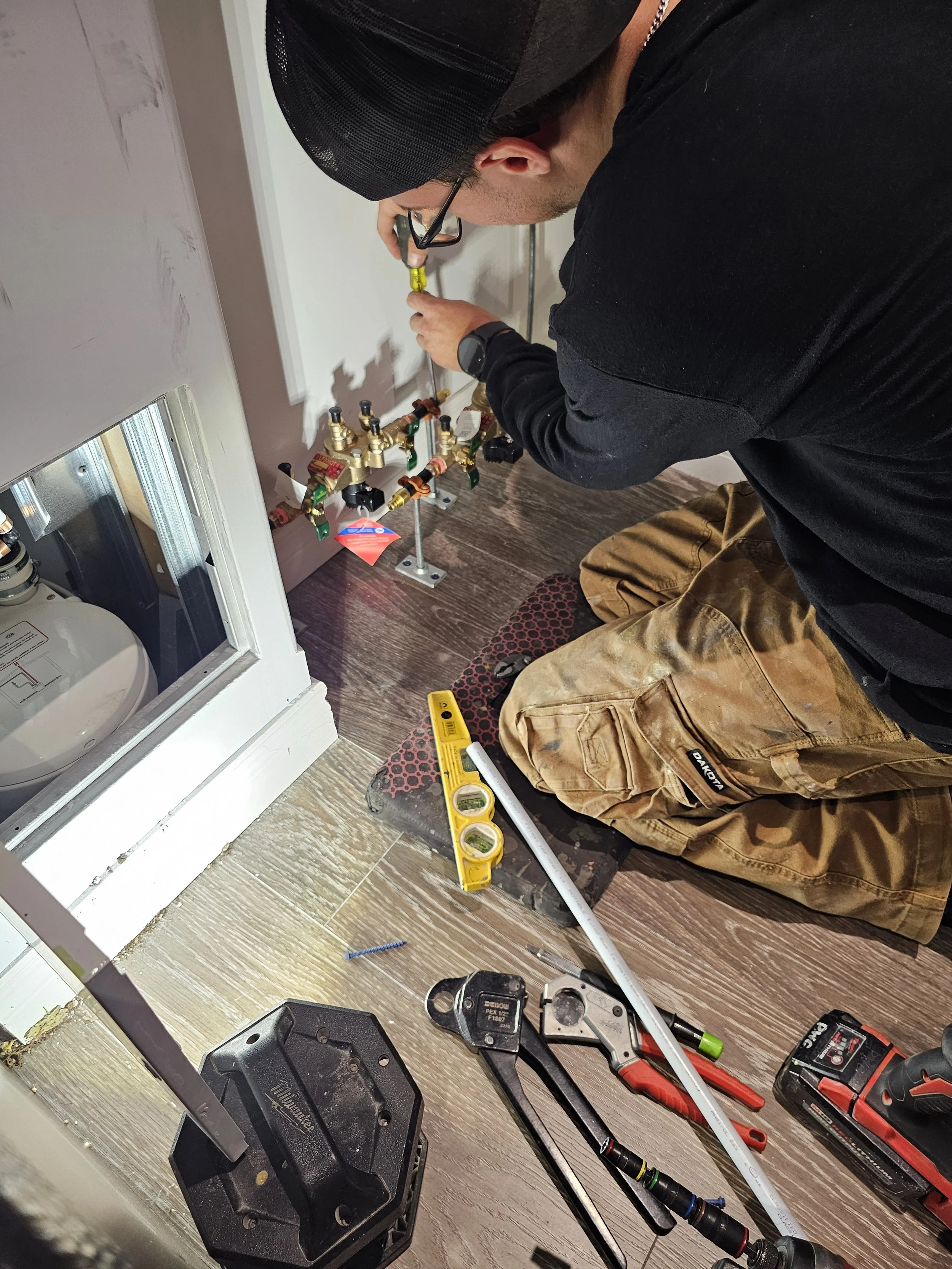 A man kneeling on the floor installing or repairing plumbing pipes, surrounded by tools such as a level, screwdrivers, a measuring tape, and a power drill.