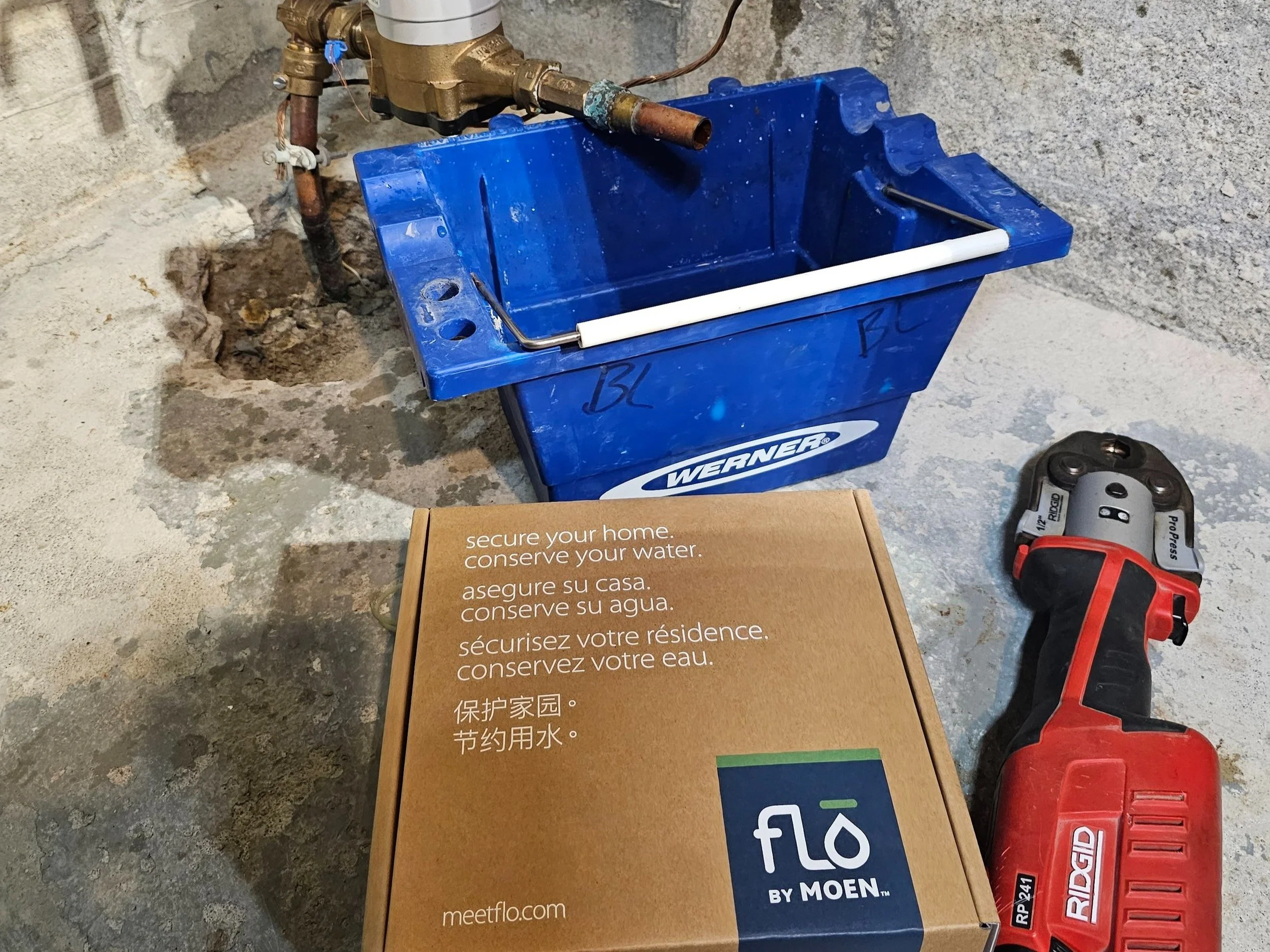 A blue Werner bucket, a cardboard box with water conservation messages in multiple languages, a red cordless drill, and plumbing pipes and valves on a concrete floor in a basement or utility room.