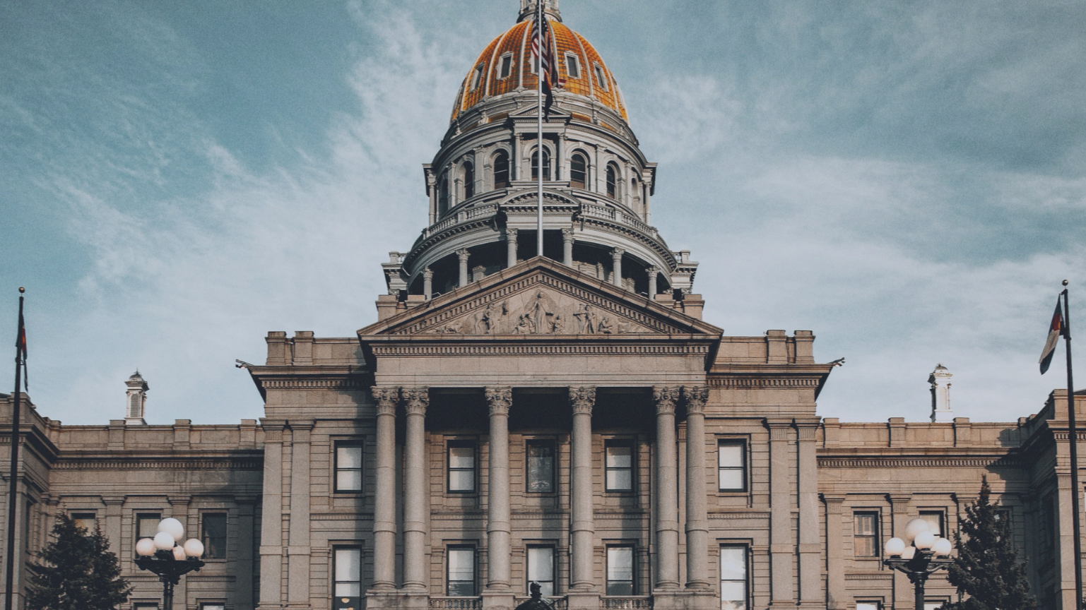 The Colorado State Capitol building with a golden dome, columns, and a historical facade, featuring flags and street lamps in front.