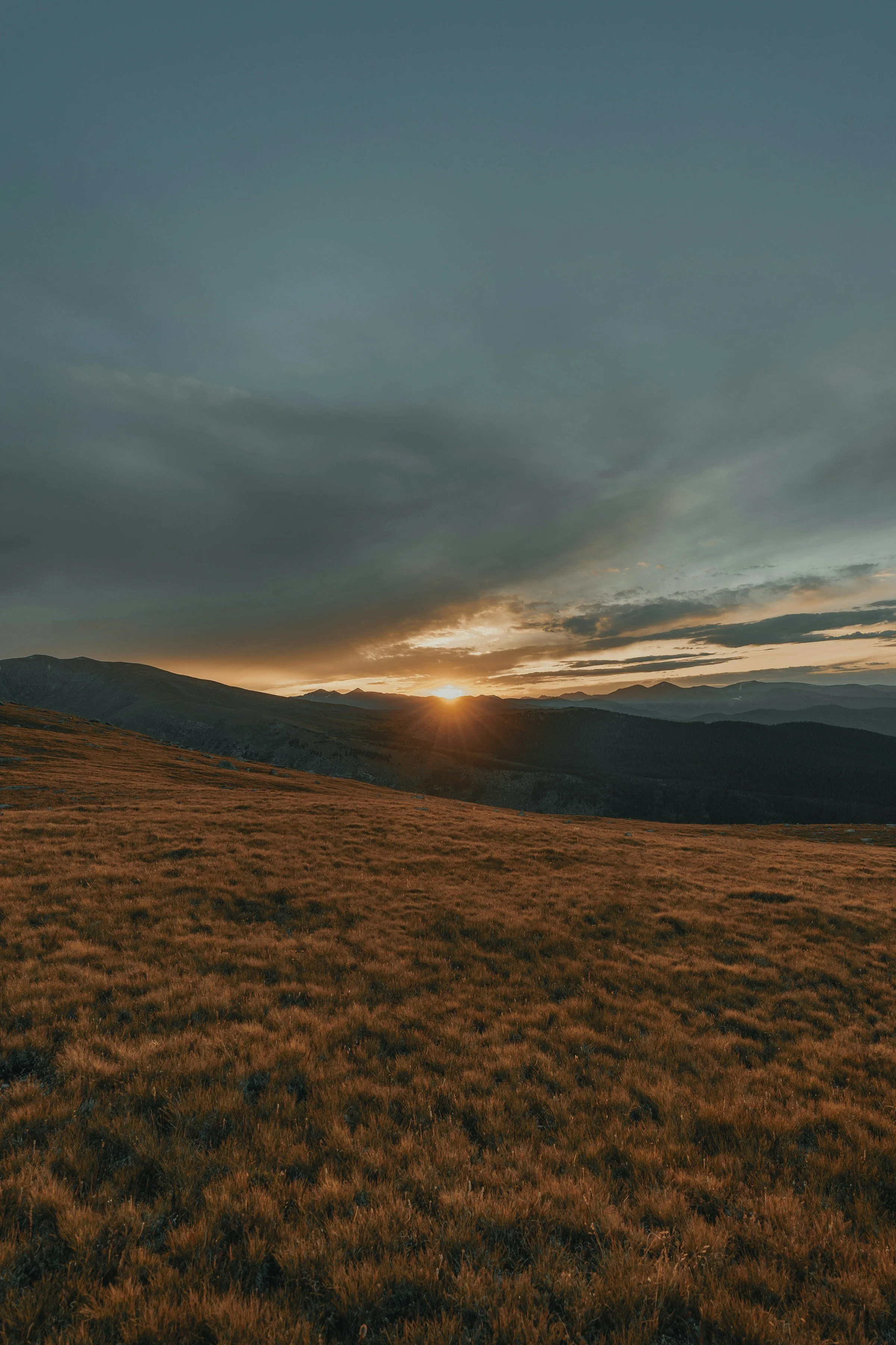 Cloudy sky above brown grassy plains with sun setting over a mountain in the horizon.