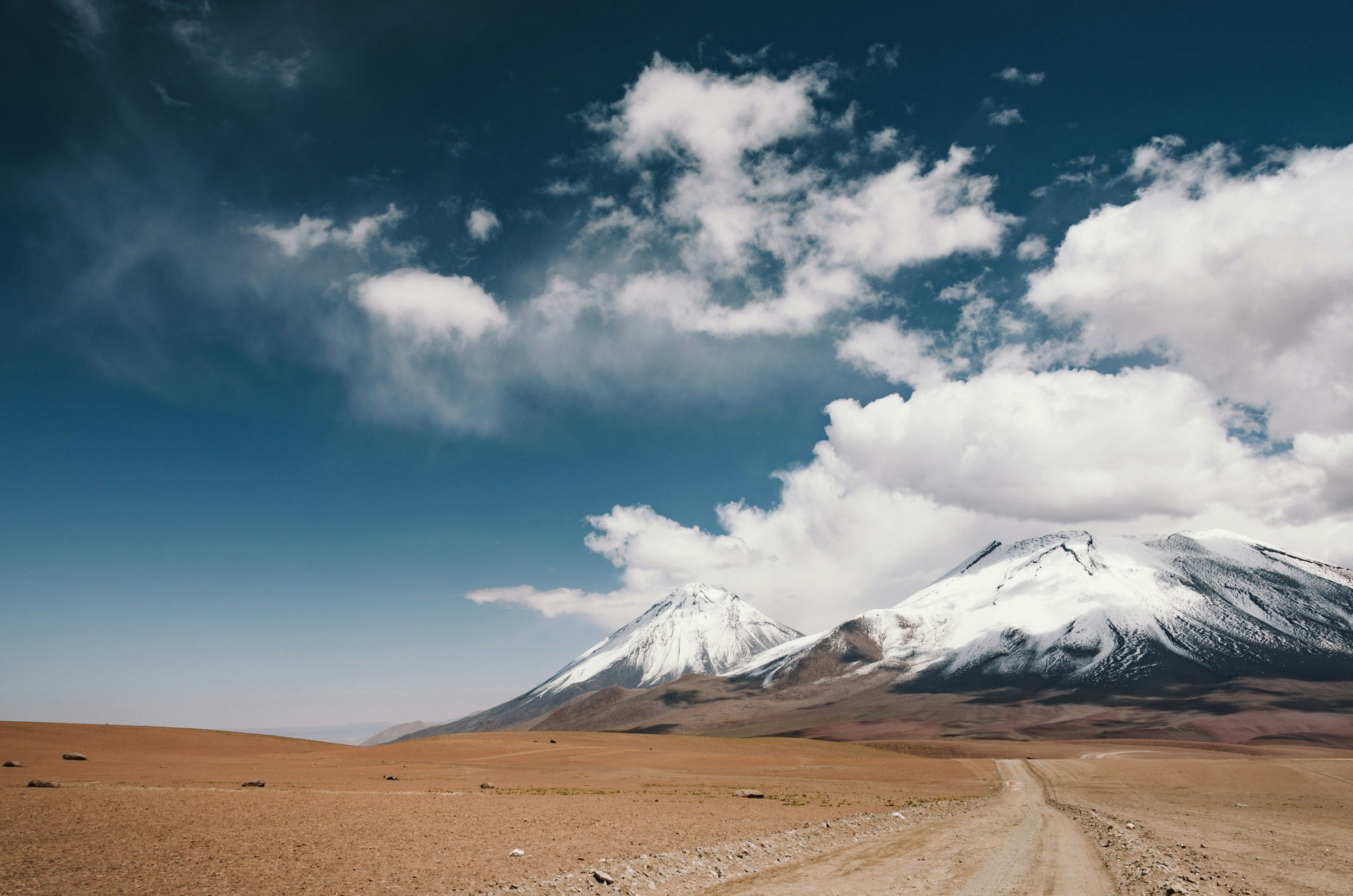 Dirt road across wide open brown plains headed toward snow capped mountain peaks in the distance.