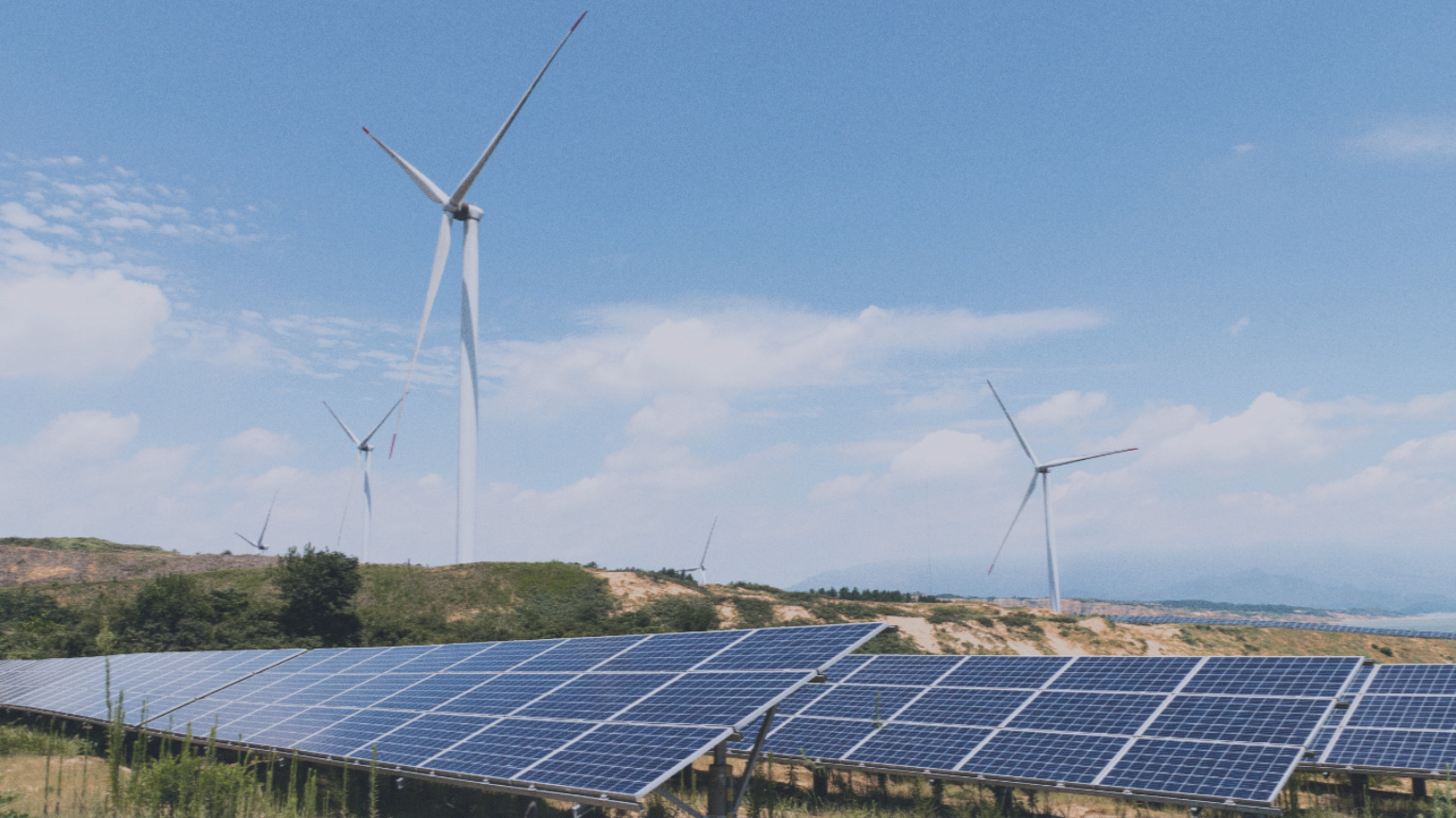 Solar panels in the foreground with wind turbines on hills in the background under a partly cloudy sky.