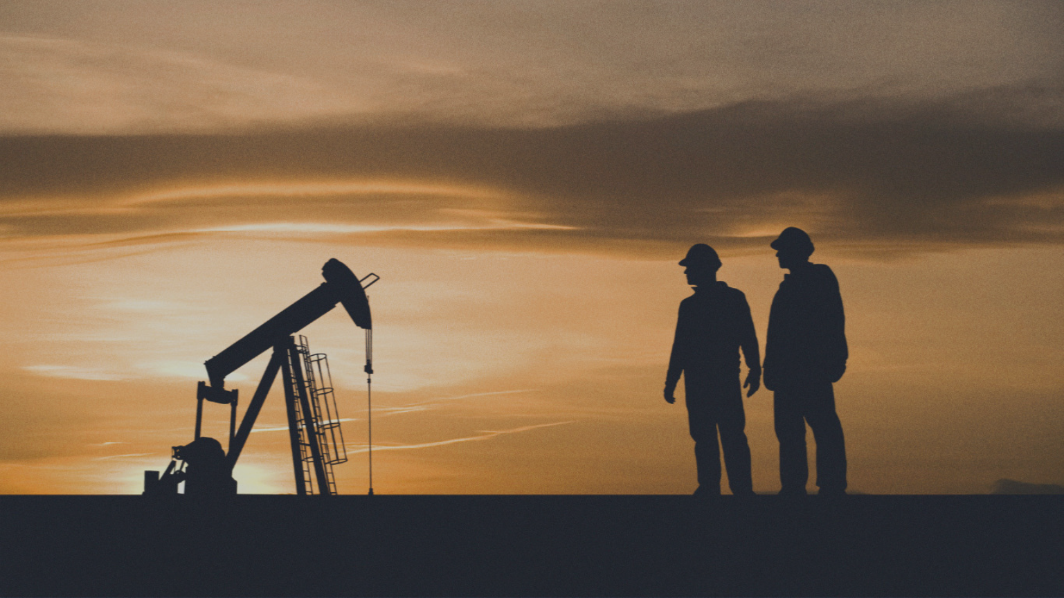 Silhouettes of two oil workers wearing helmets standing near an oil pumpjack during sunset.