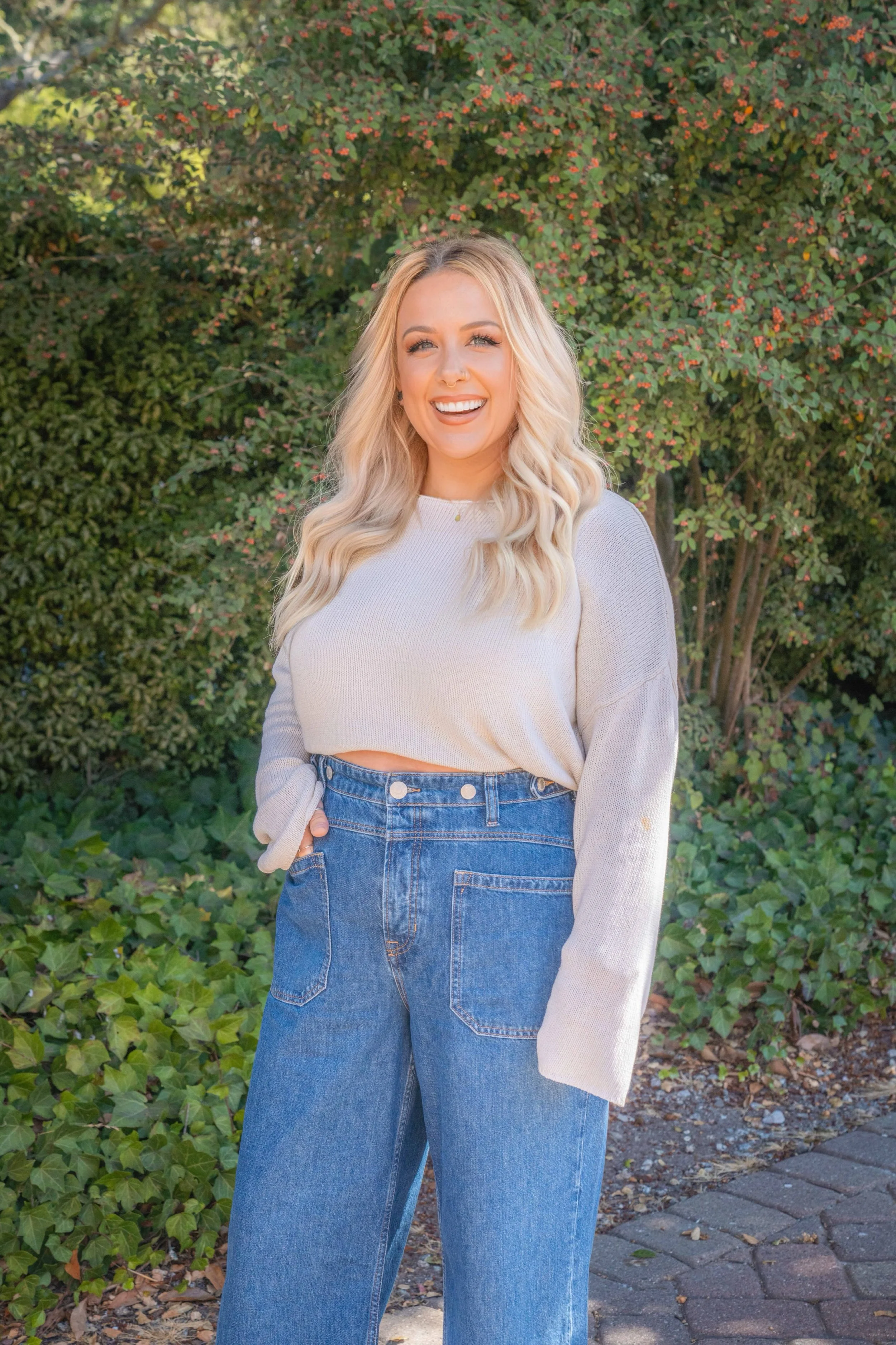 A smiling young Caucasian woman with long, wavy blonde hair standing outdoors in front of green bushes with red berries, wearing a beige cropped sweater and high-waisted blue jeans.