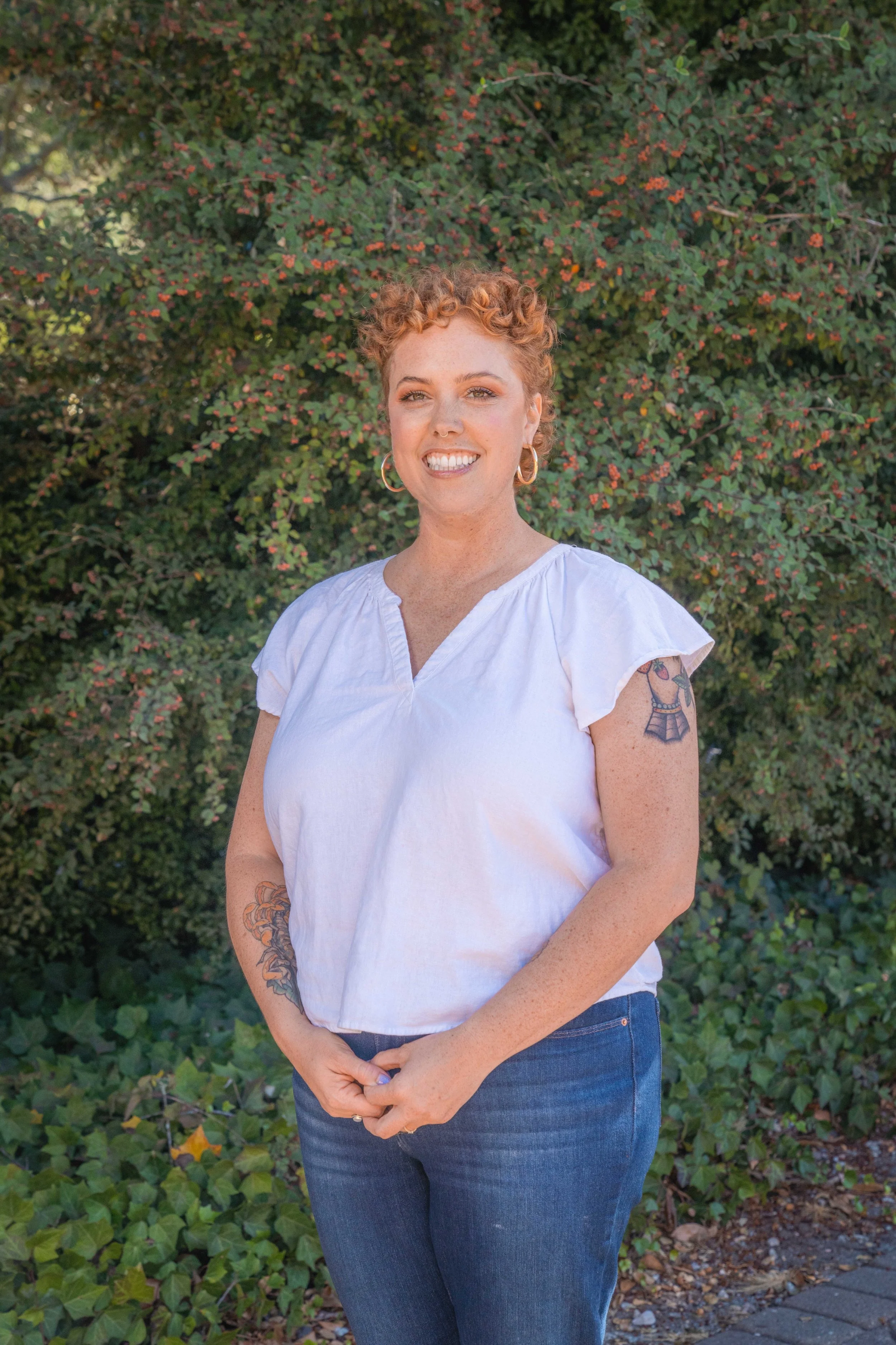 A woman with short curly red hair, wearing hoop earrings and a white blouse, standing outdoors in front of green foliage with small pink flowers.