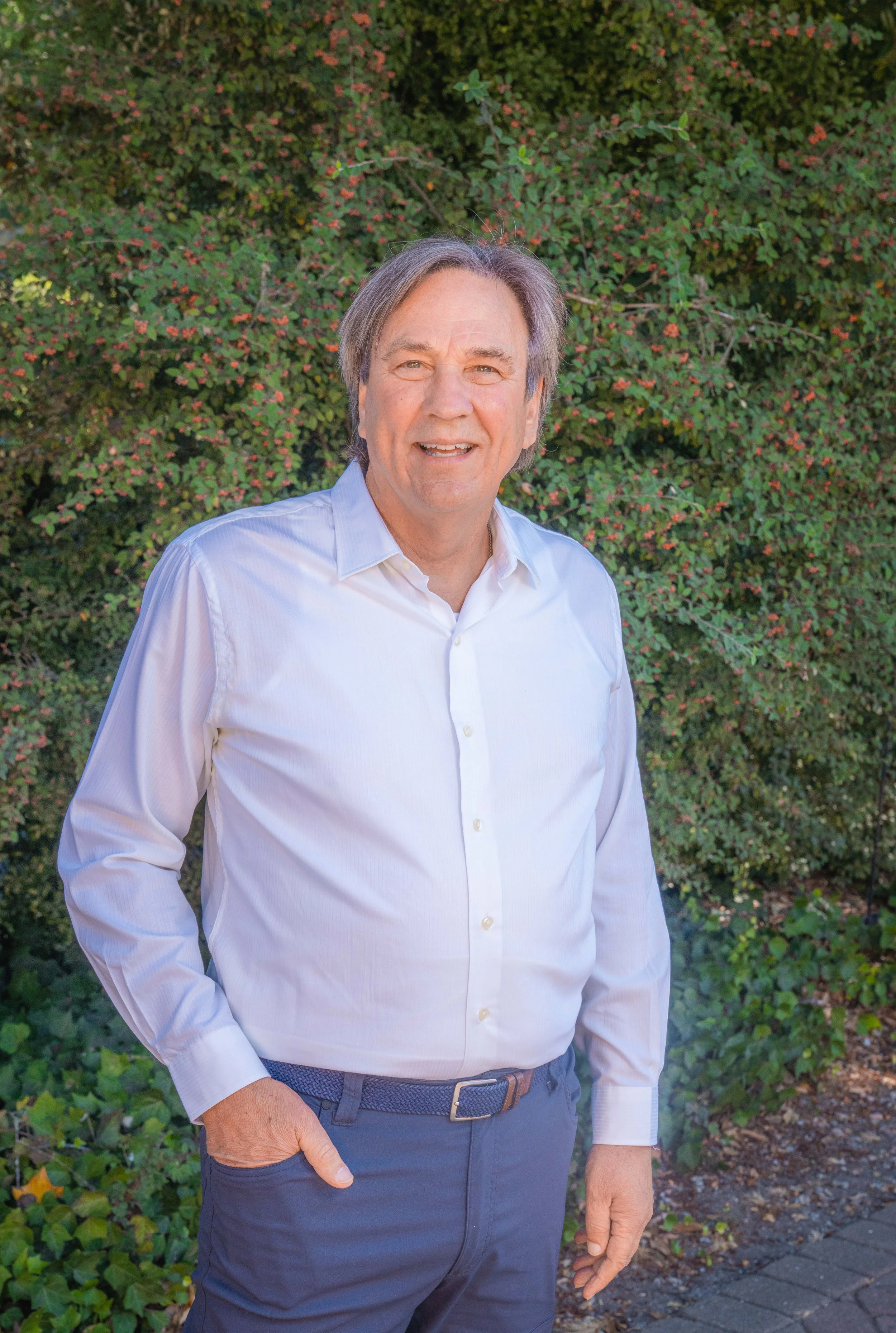 A middle-aged man with gray hair, wearing a white button-up shirt and dark blue pants, standing outdoors in front of green bushes with small pink flowers, smiling at the camera.