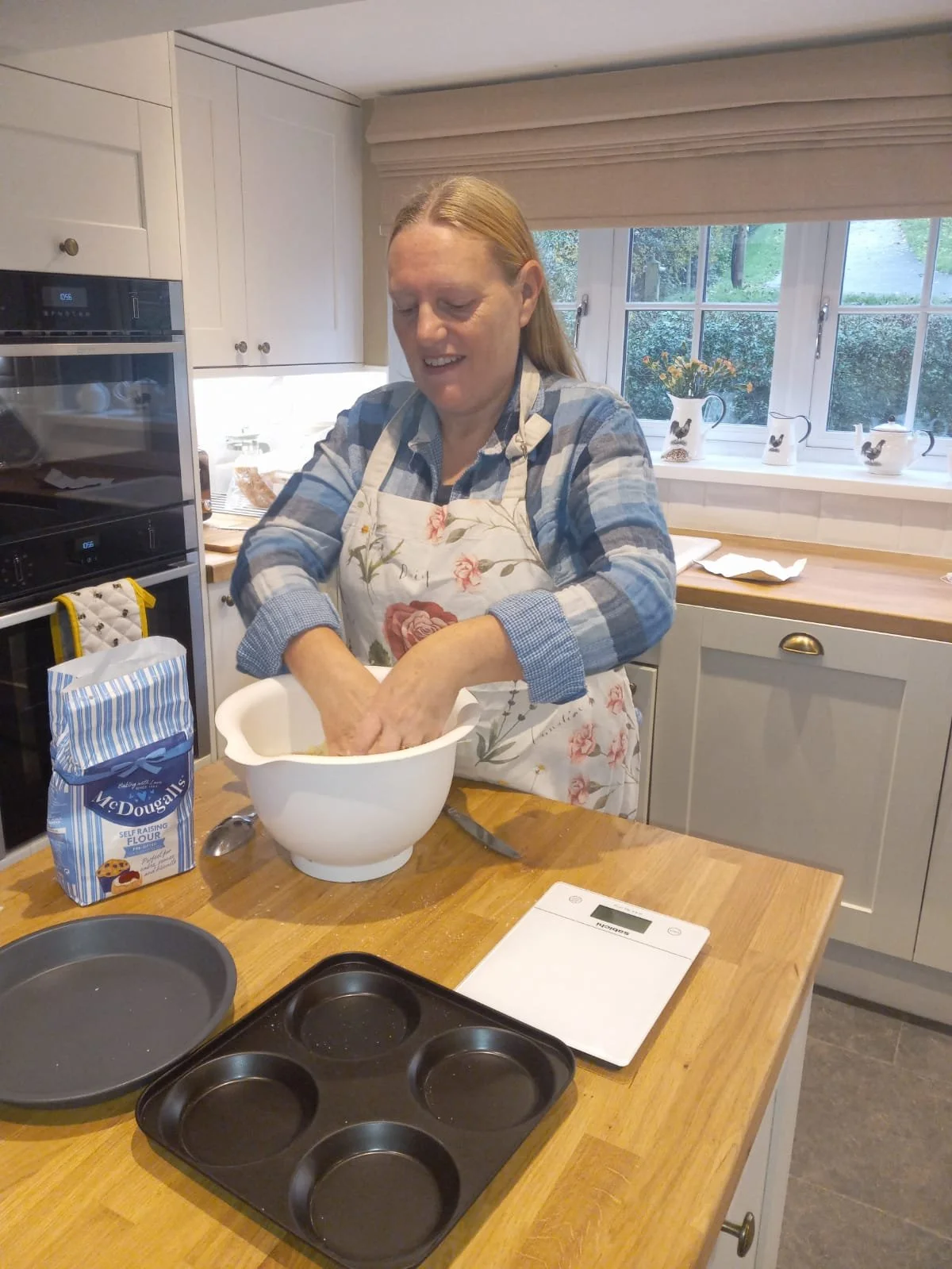 A woman wearing a floral apron and a checkered shirt is mixing ingredients in a white bowl in a cozy kitchen with white cabinets, a window with decorative mugs, and a wooden countertop.