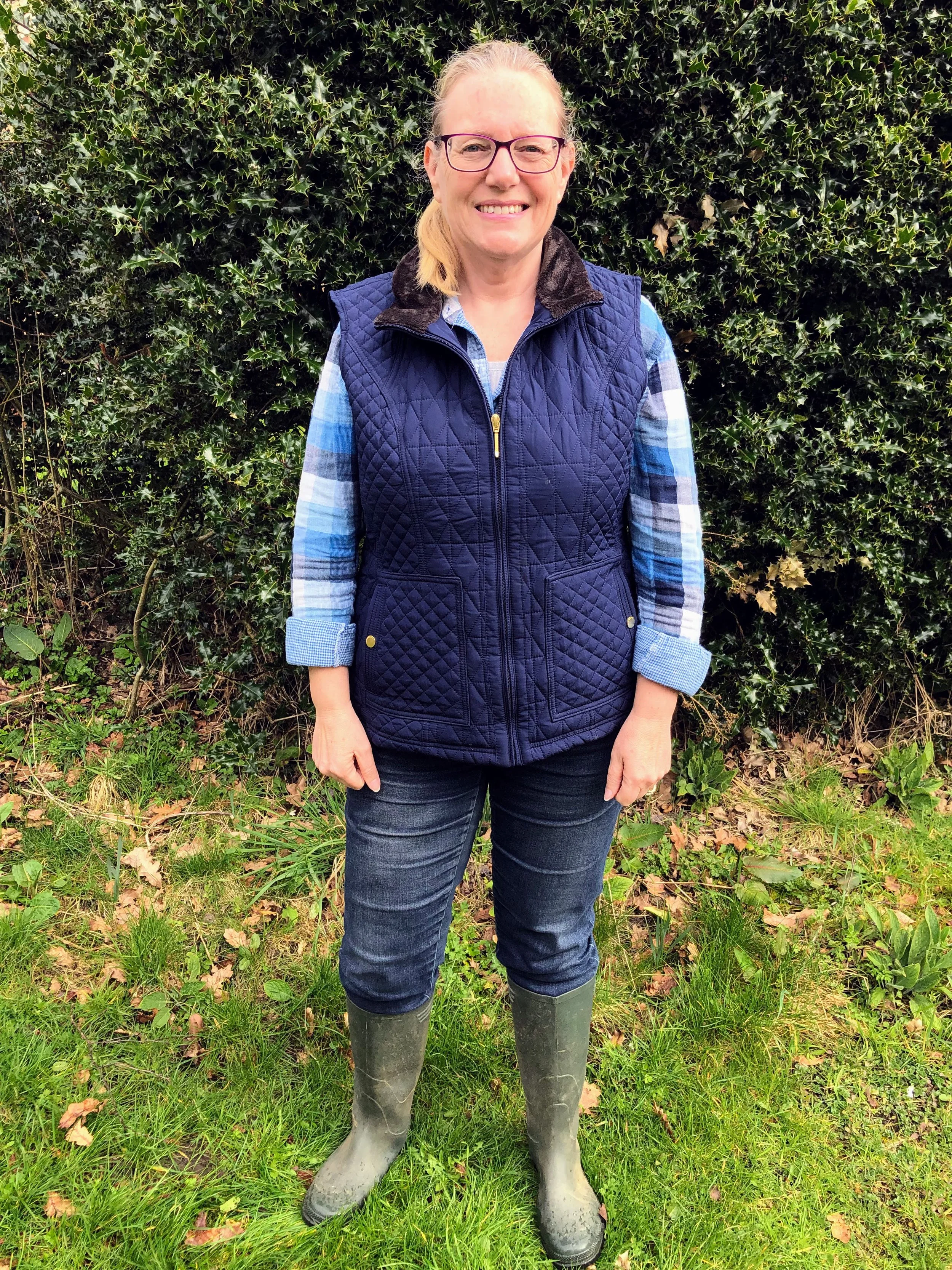 Tina Jesson standing outdoors in front of a dense hedge of holly leaves, wearing glasses, a blue plaid shirt, a navy quilted jacket, jeans, and tall green wellington boots.