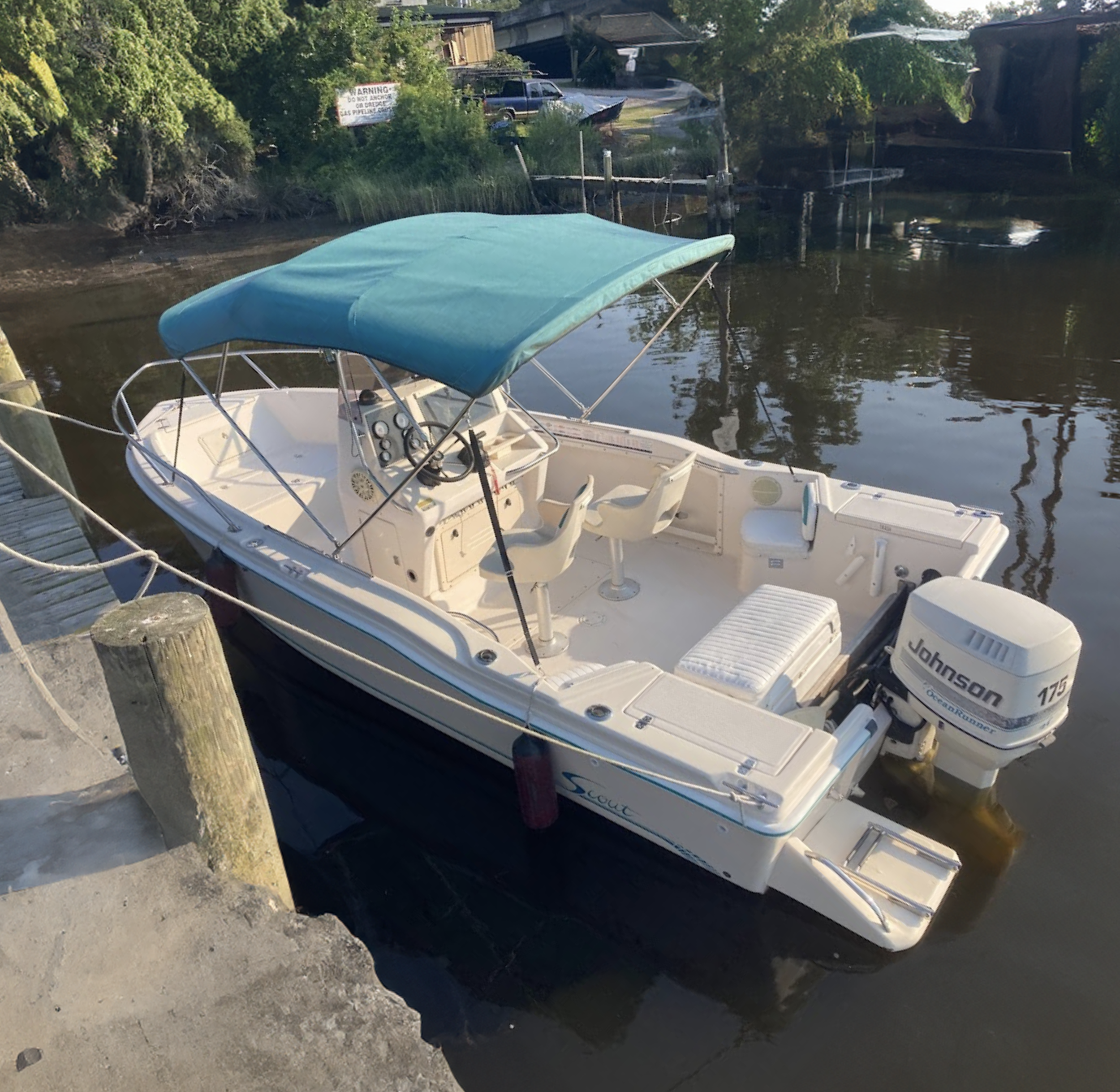 A small motorboat docked at a river, featuring a blue canopy, two seats, and a Johnson 175 outboard engine.