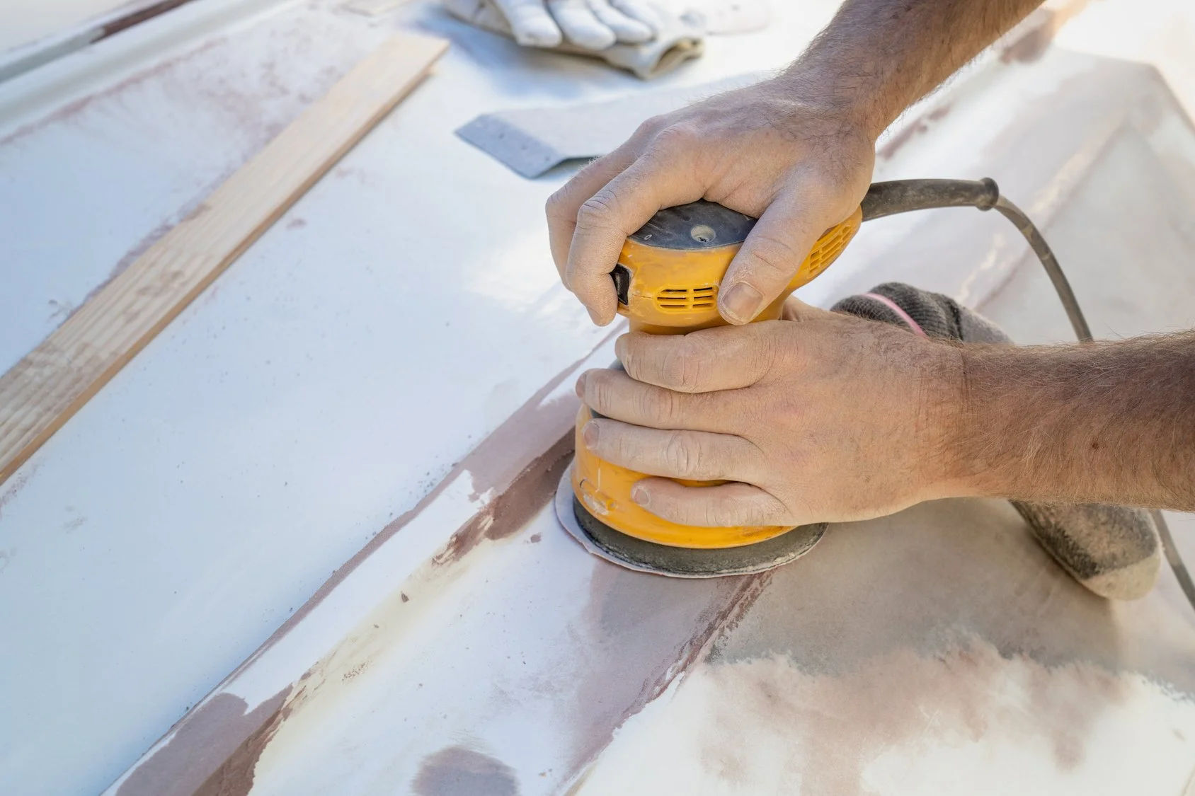 Close-up of hands using a yellow electric sander on wood surface, with dust and a wooden plank nearby.