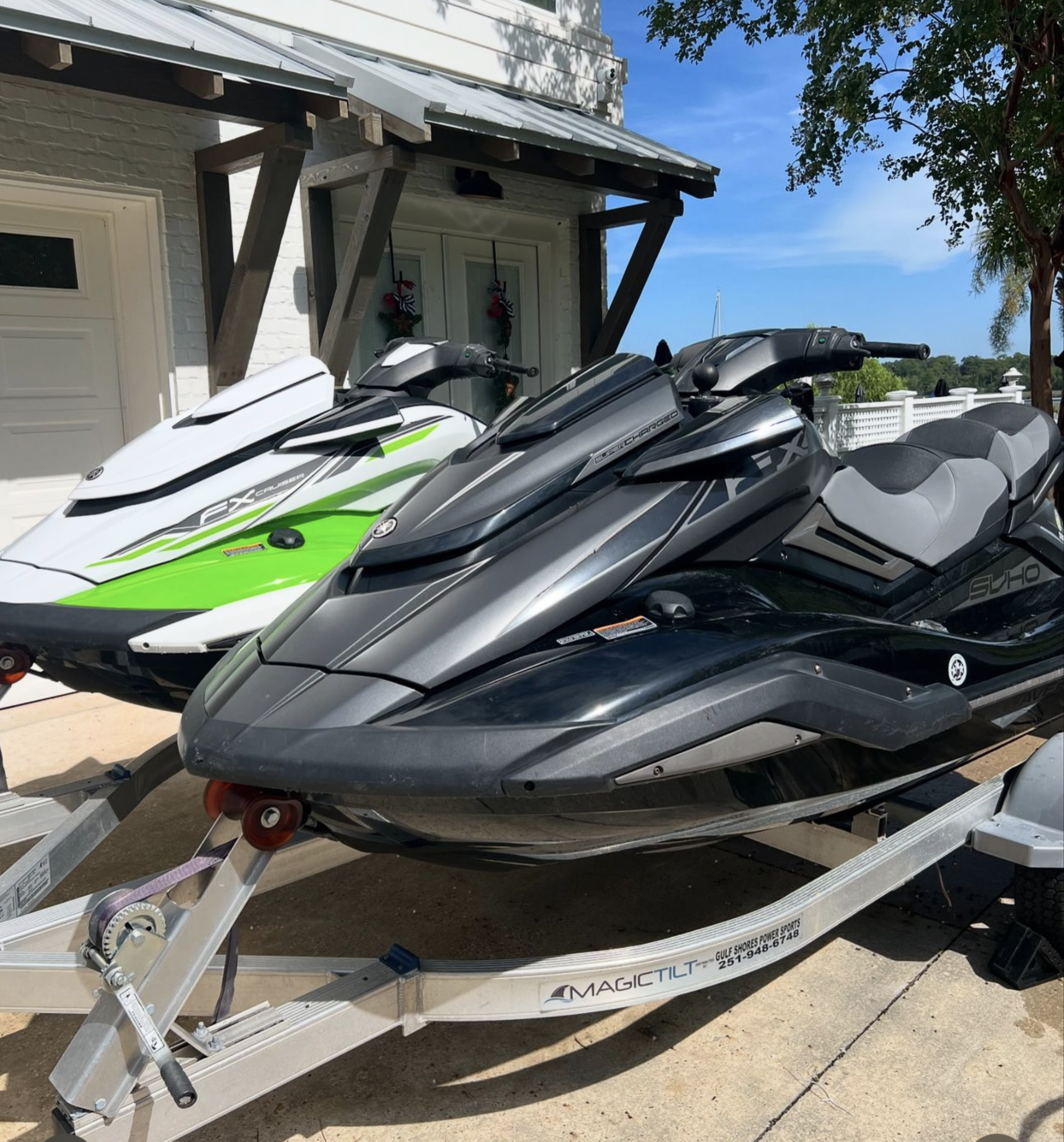 Two personal watercraft, one white and green and the other black and gray, are parked on a trailer in front of a building with a wood beam roof.