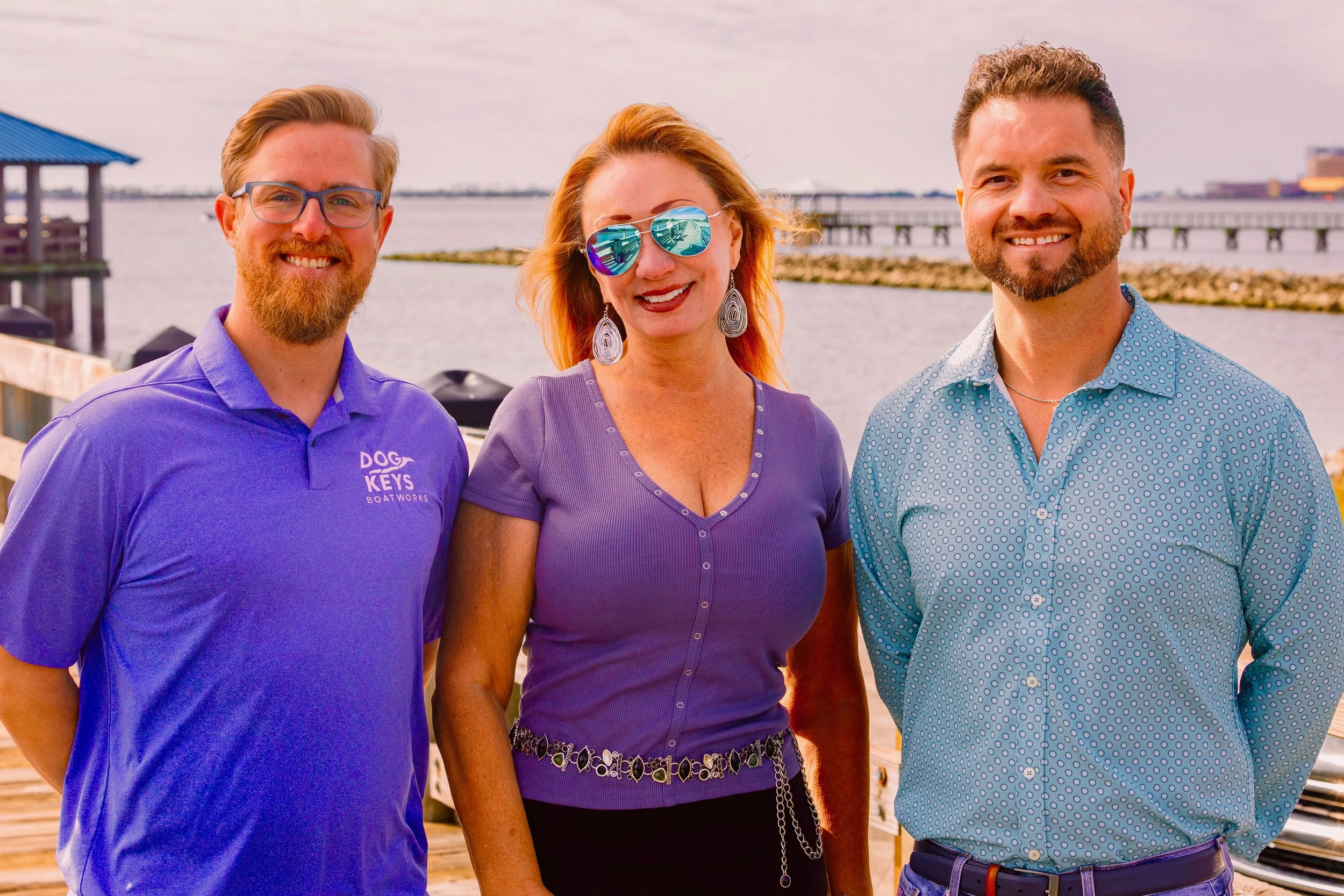 Three people smiling outdoors, standing by a waterfront, wearing casual clothes including a purple shirt, a lilac top, and a teal shirt. The background features water, a pier, and buildings on the horizon.