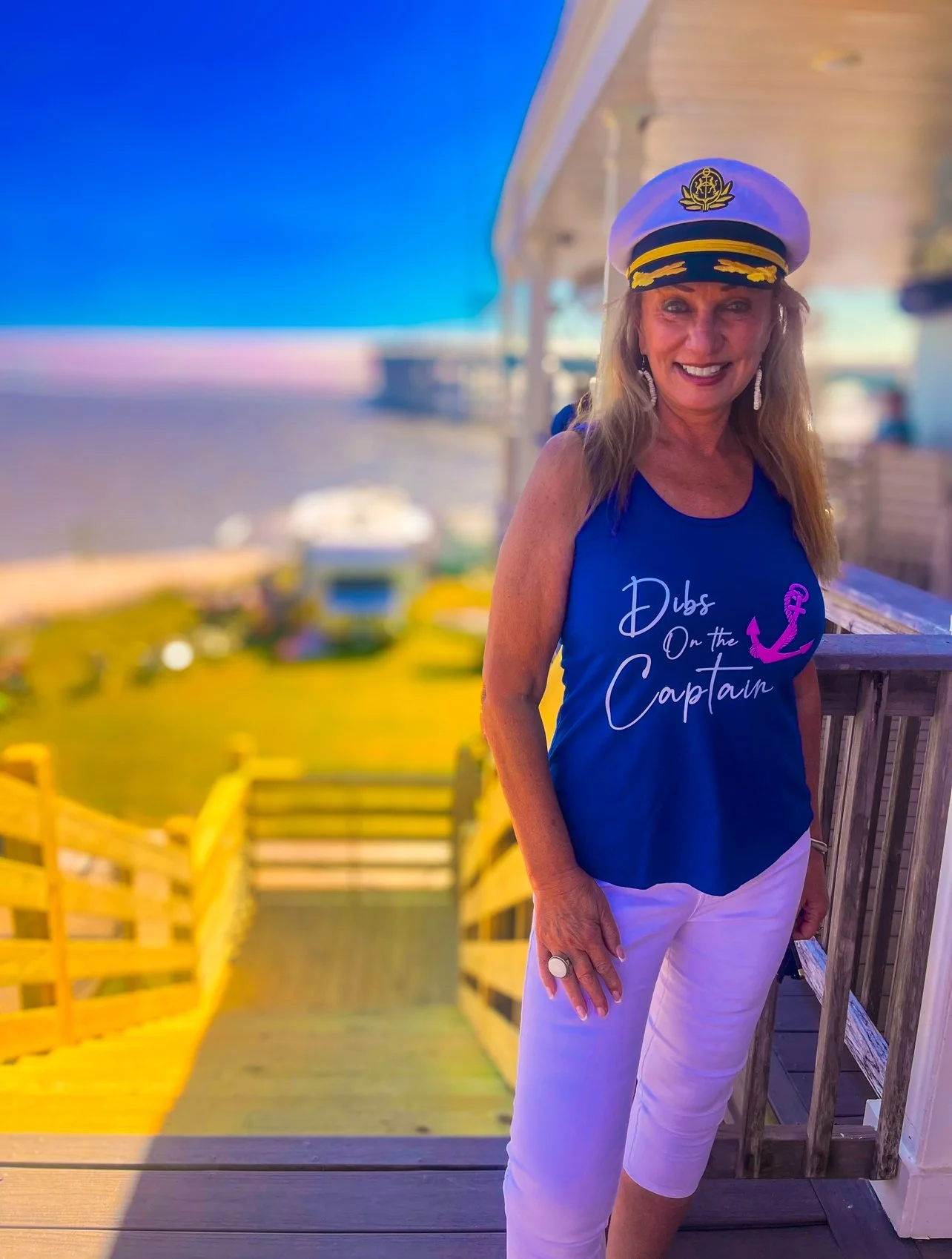 Woman wearing a captain's hat, a blue tank top with "Dibs on the Captain" text, and white pants, standing on a wooden deck with a beachfront view in the background.