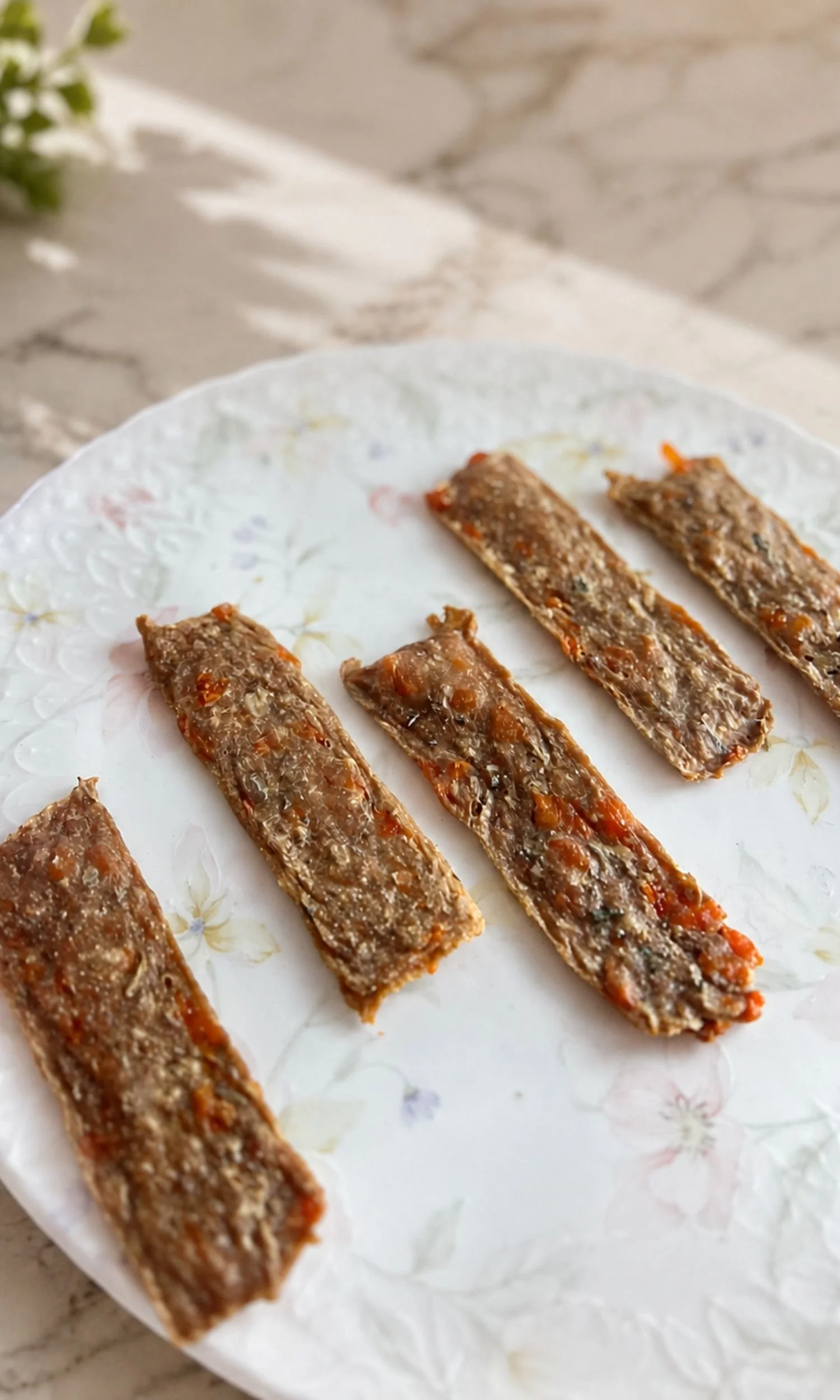 Several rectangular pieces of homemade jerky dog treats on a white floral plate, set on a marble countertop.