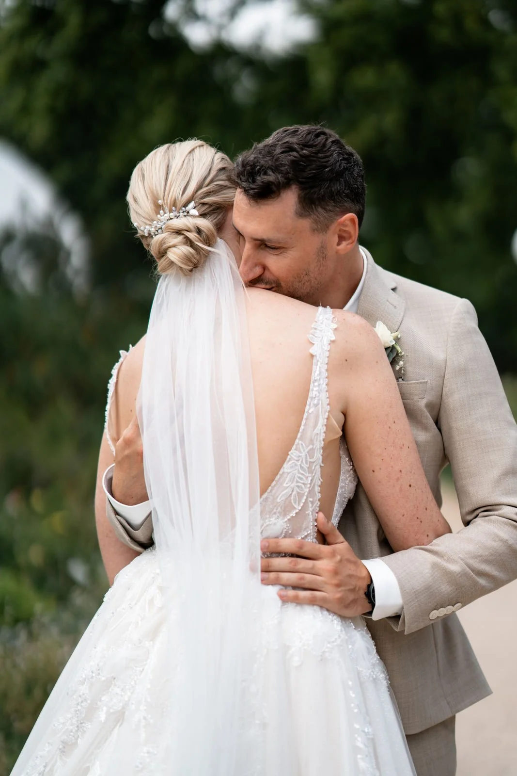 Ein Brautpaar umarmt sich liebevoll bei einer Hochzeit im Freien, die Braut trägt ein weißes Kleid mit Spitze und einen Schleier, der Bräutigam trägt einen hellbeigen Anzug.
