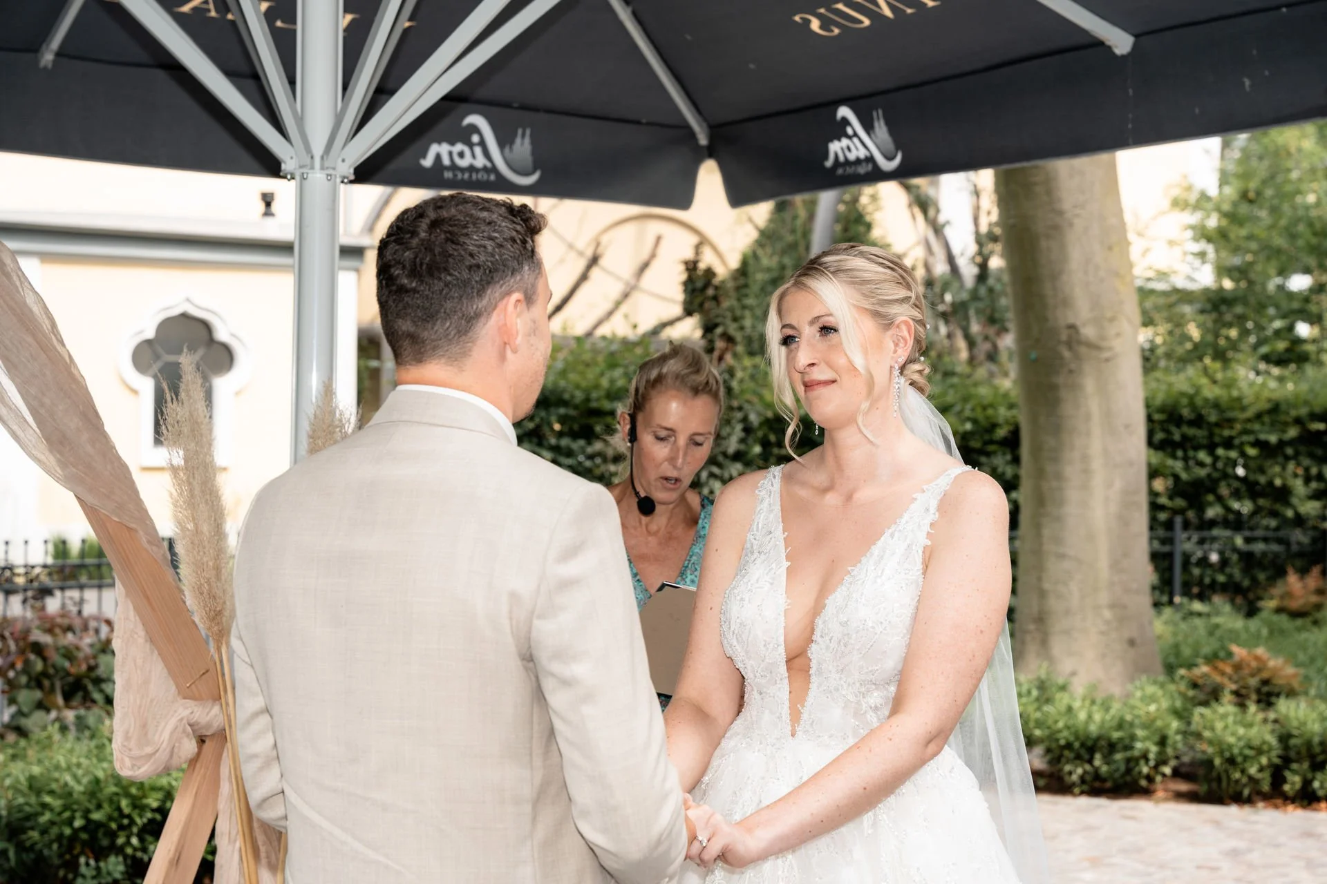 Braut und Bräutigam bei ihrer standesamtlichen Hochzeit im Freien, die Braut trägt ein weißes Hochzeitskleid mit tiefem Ausschnitt, die Trauung findet unter einem großen schwarzen Schirm statt.