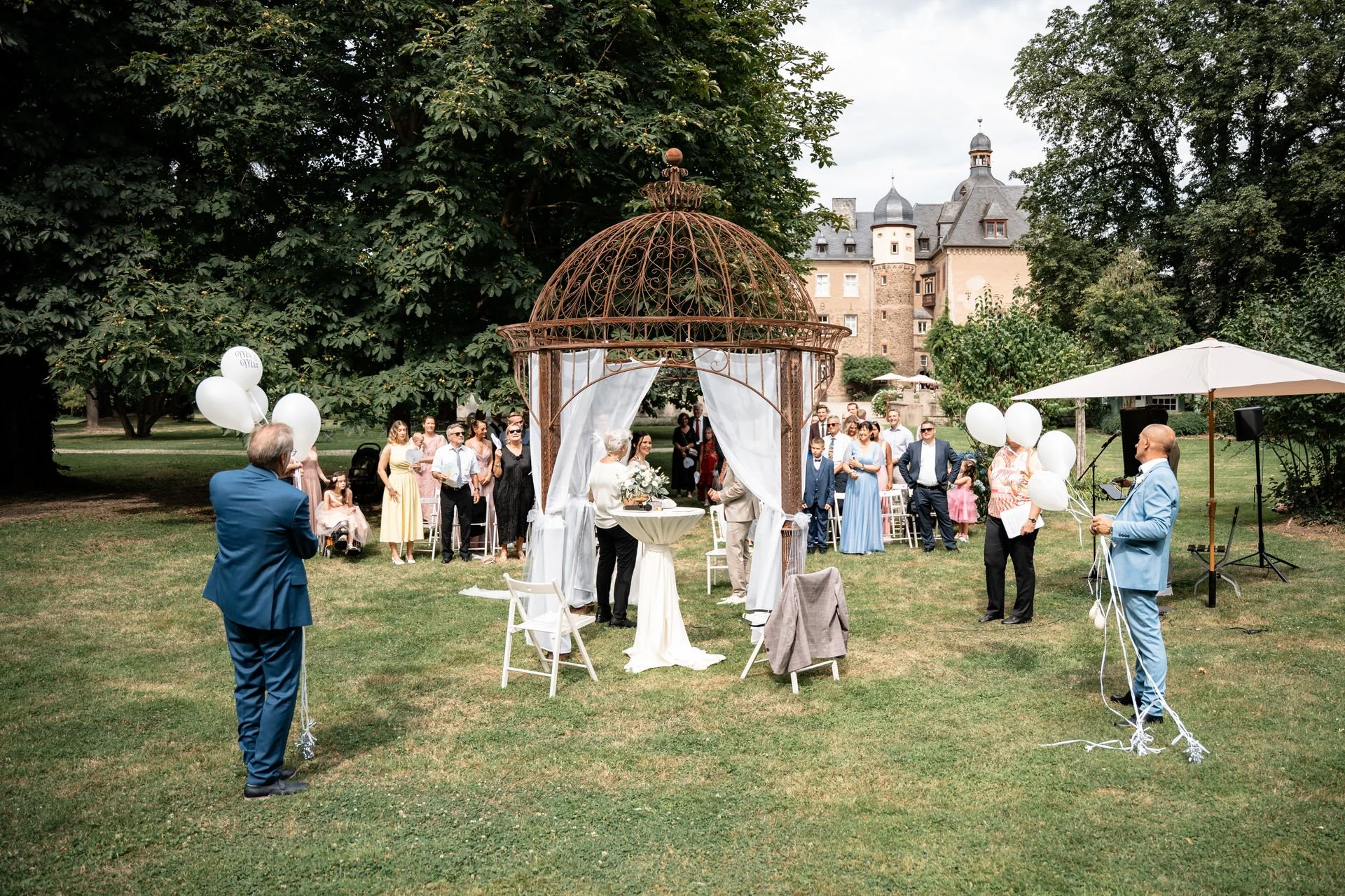 Eine Hochzeit im Freien mit Gästen, die vor einer rustikalen Pavillon im Garten stehen. Es gibt weiße Ballons und eine historische Burg im Hintergrund.