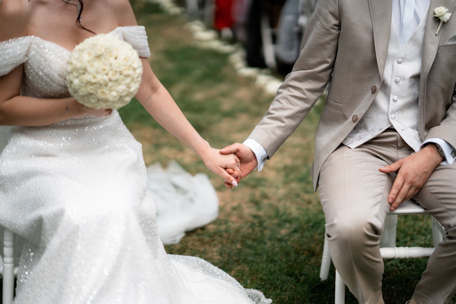 Ein Brautpaar hält sich bei ihrer Hochzeitszeremonie an den Händen, die Braut trägt ein weißes, funkelndes Hochzeitskleid und hält einen Blumenstrauß, während der Bräutigam in einem beige Anzug sitzt.