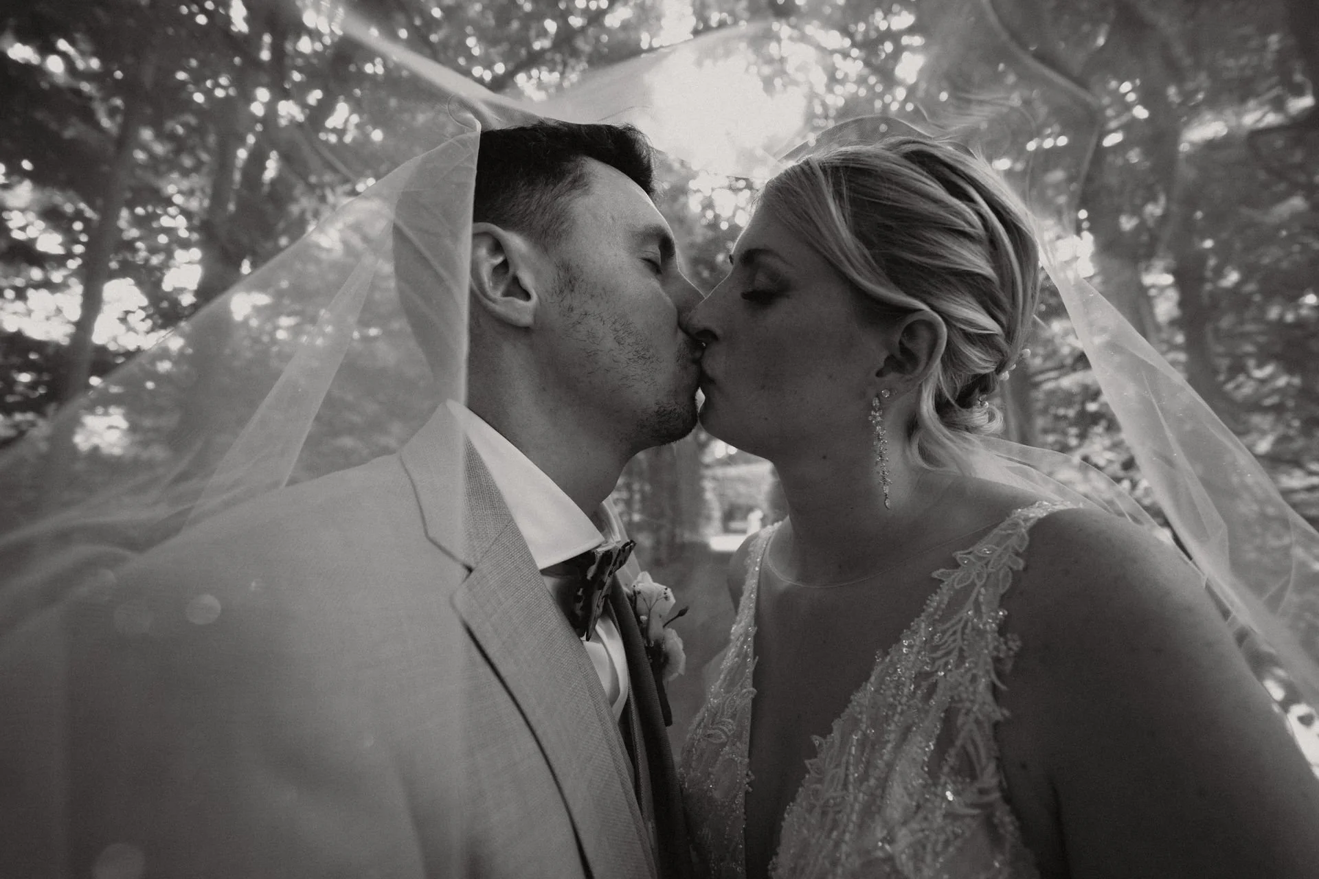 Braut und Bräutigam küssen sich unter einem Schleier im Freien, mit Bäumen im Hintergrund