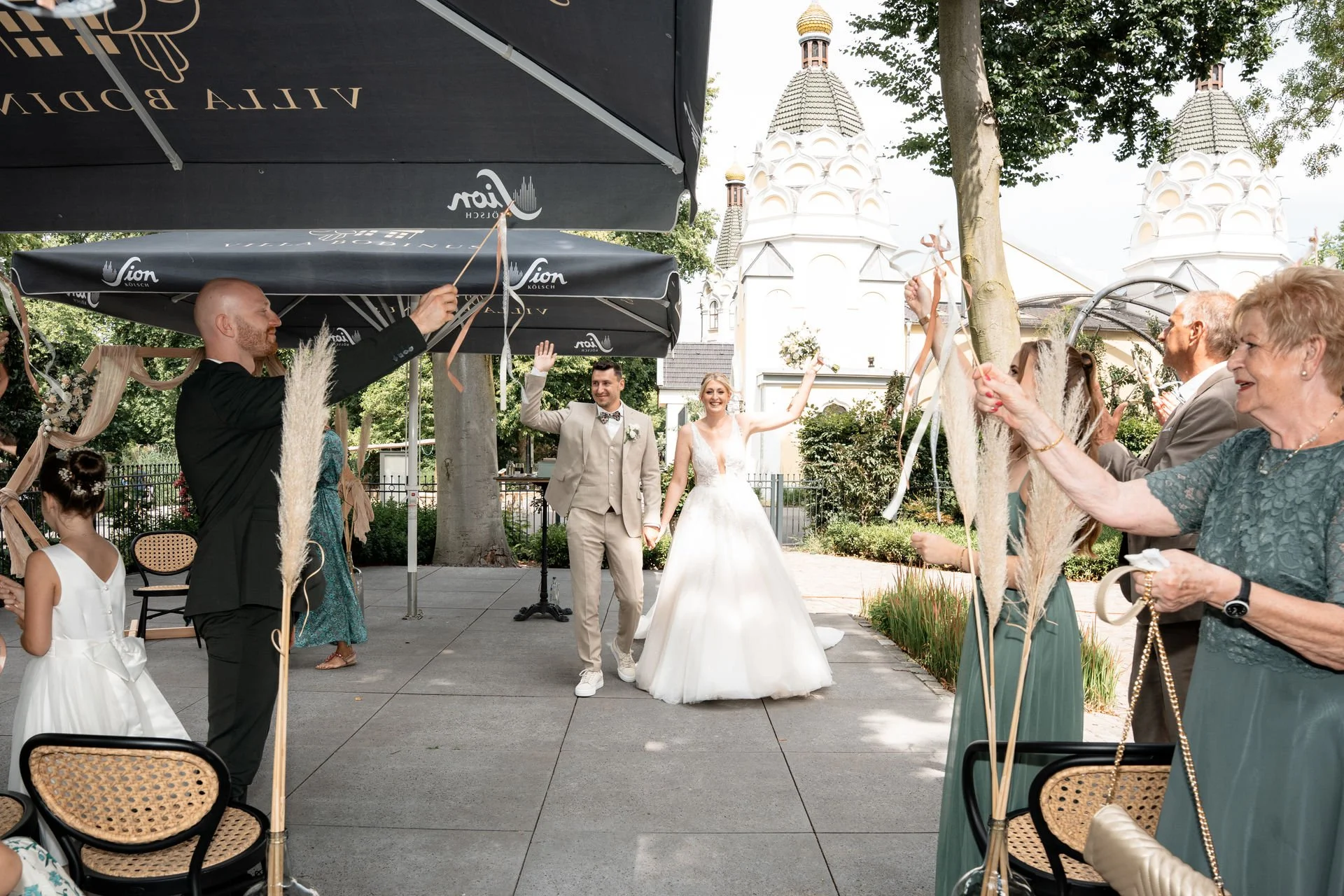 Ein frisch verheiratetes Paar hält Händchen und läuft durch einen Garten, umgeben von Freunden und Familienmitgliedern, die ihnen mit schön gestalteten Blumen und Zelten gratulieren.
