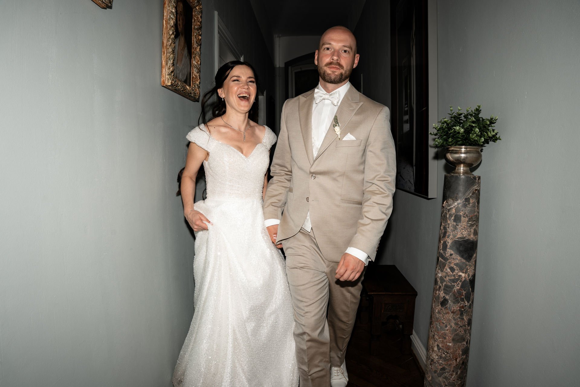 Ein Paar in Hochzeitskleidung geht im Flur eines Hauses. Das Brautpaar lacht und scheint fröhlich zu sein.