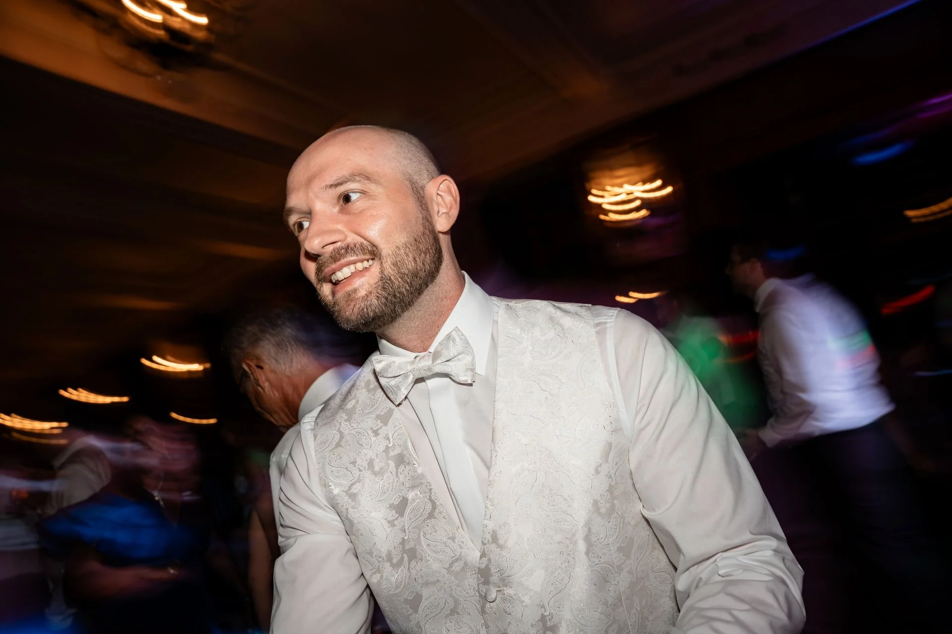Ein Mann in einem weißen Anzug mit Fliege, der auf einer Party tanzt und lächelt, im Hintergrund verschwommene Lichter und andere Gäste.