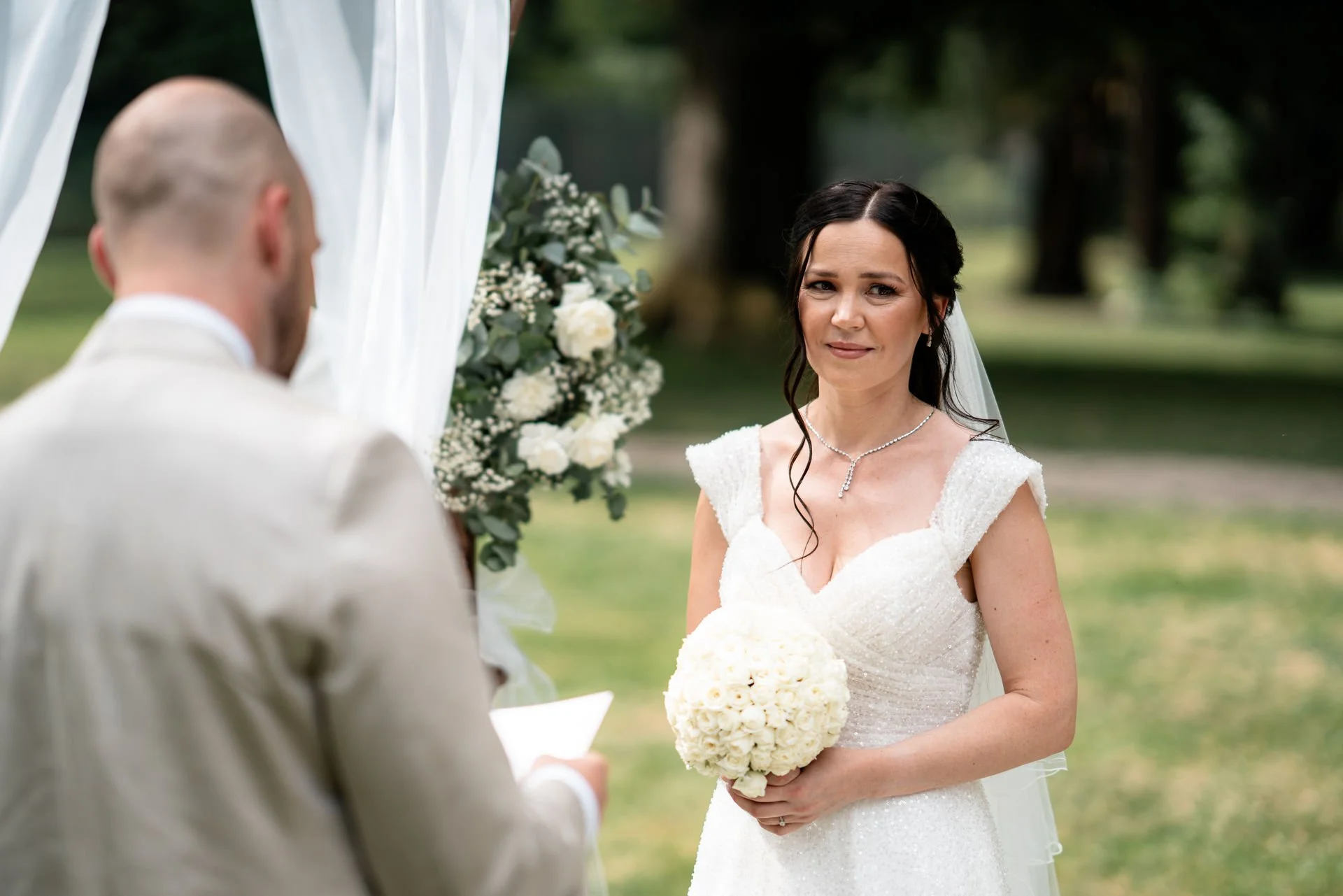 Braut hält einen Blumenstrauß bei einer Hochzeitszeremonie im Freien, während der Bräutigam, der nur von hinten sichtbar ist, ihr gegenübersitzt.