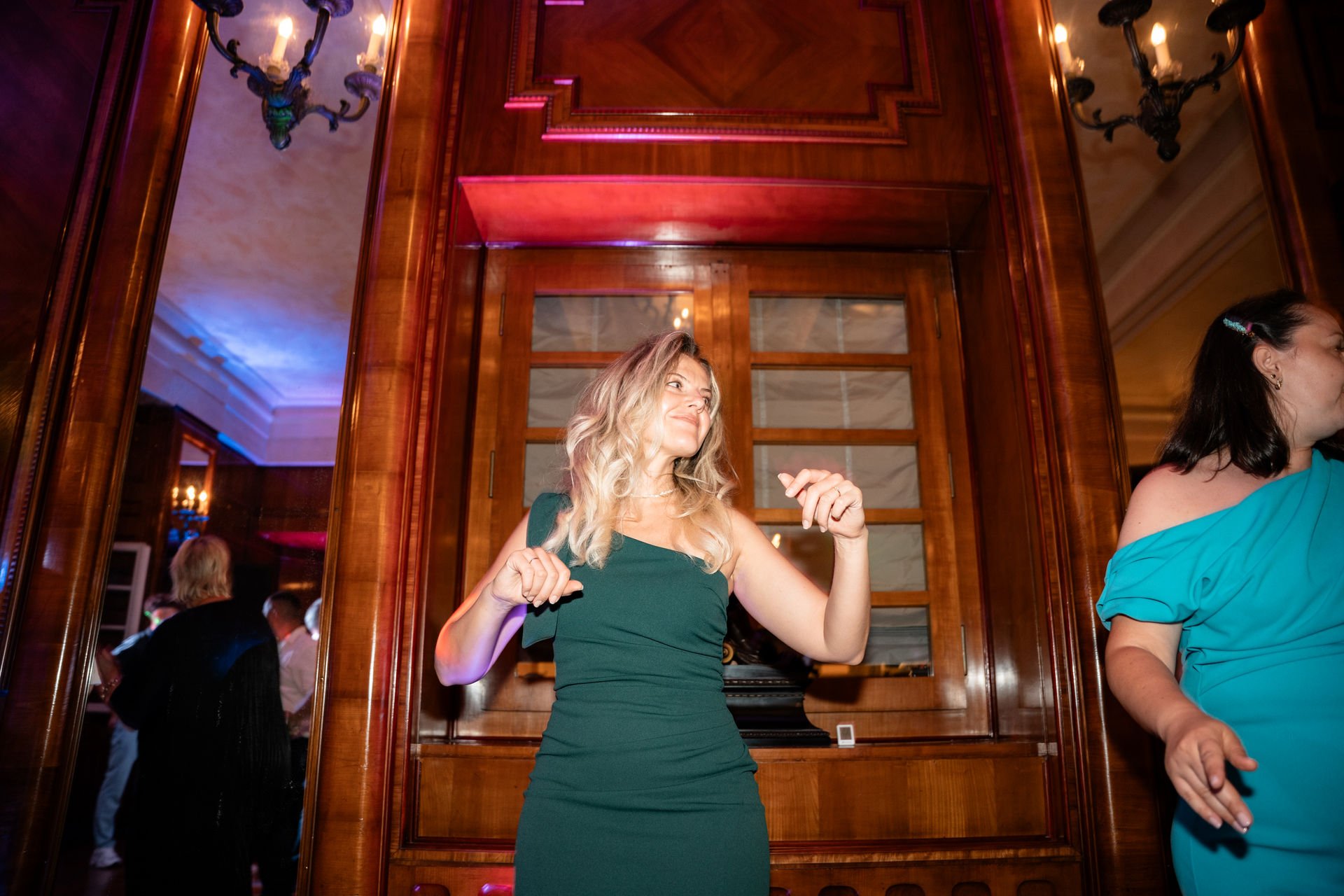 Eine lachende rote Haar Frau in einem dunkelgrünen Abendkleid tanzt in einem elegant eingerichteten Raum mit Holzpaneelen und Kronleuchtern.