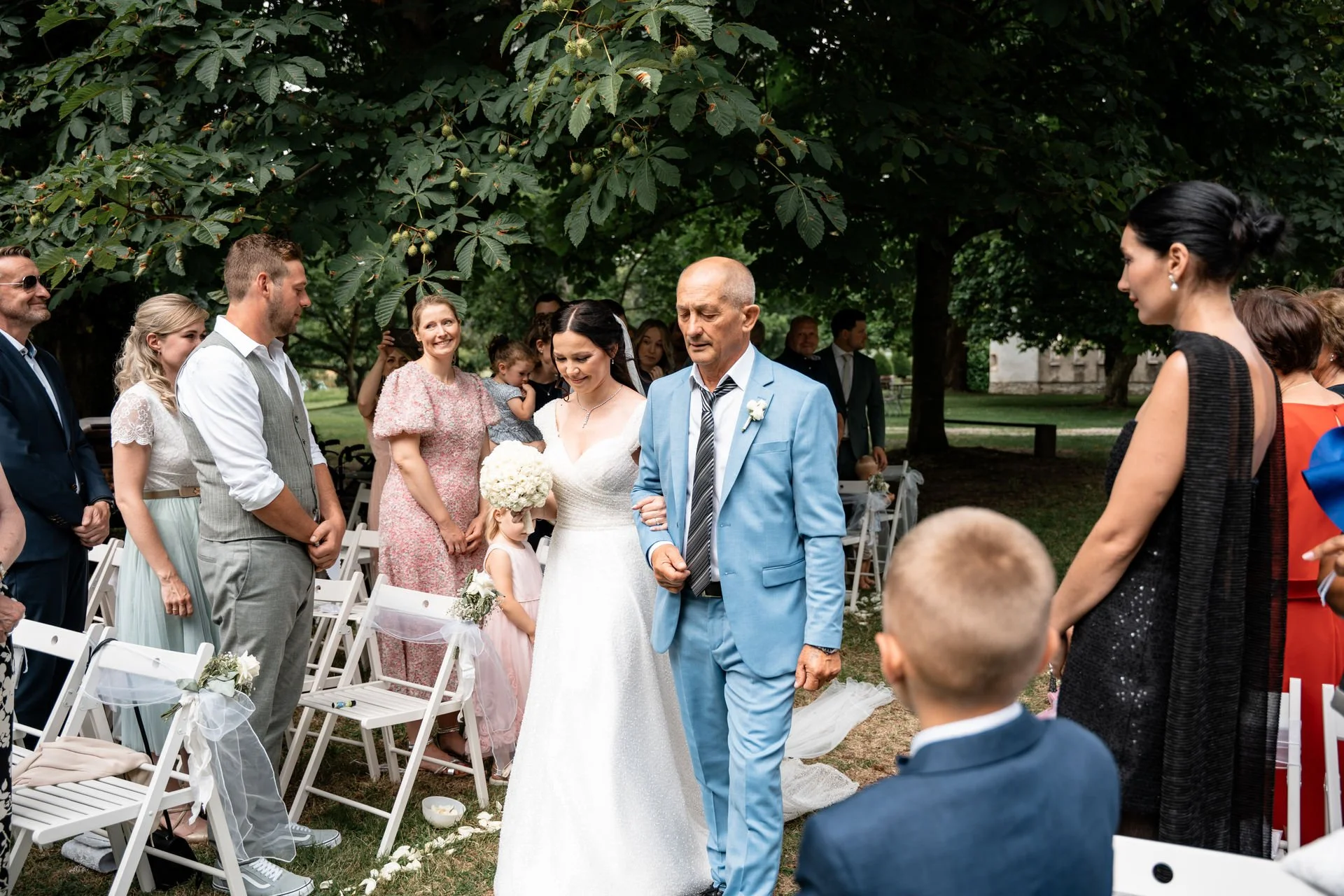 Hochzeit im Freien mit Braut in weißem Kleid, die von einem älteren Mann in blauer Jacke begleitet wird, umgeben von Gästen in eleganten Kleidern und Anzügen, im Schatten großer Bäume.