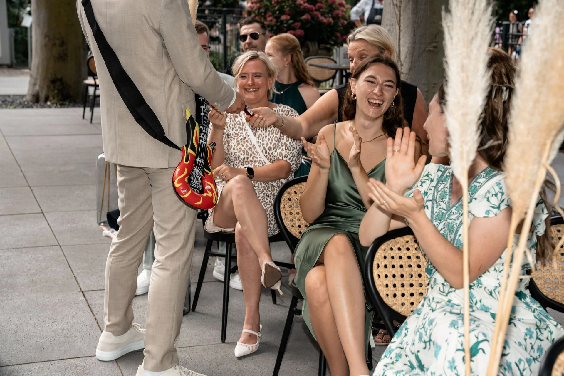 Menschen beim Treffen im Freien, lachend und im Gespräch, eine Frau erhält ein Geschenk, eine Frau trägt ein Sommerkleid mit Papageienmotiv, im Hintergrund Bäume und weitere Personen.