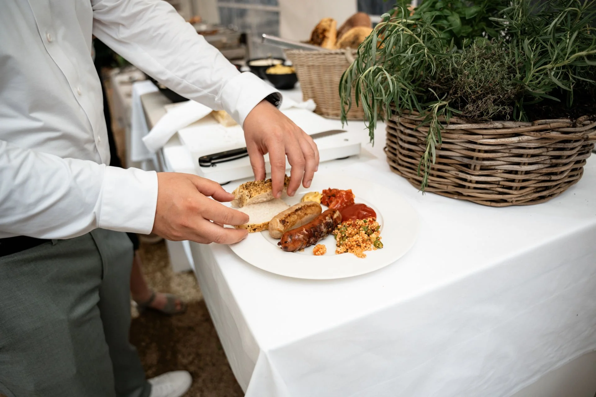 Person legt Brot auf einen Teller mit Würstchen, Tomatensauce, Couscous und Senf, auf einem weißen Tisch mit Pflanzen und Essen im Hintergrund.