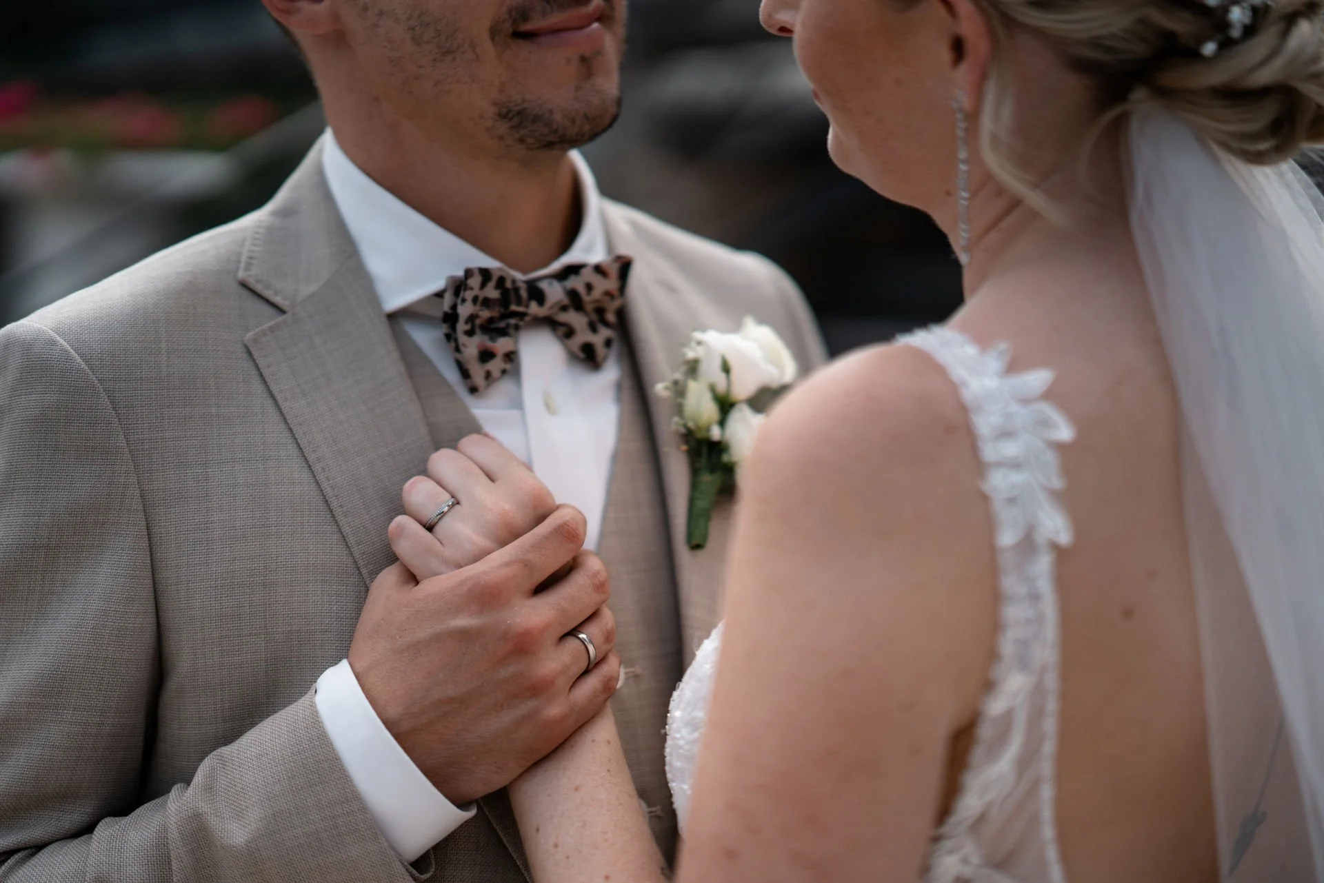 Nahaufnahme eines Paares bei der Hochzeit, das sich gegenübersteht, sie hält seine Hand, er trägt einen beige Anzug mit floral gemustertem Fliege, sie trägt ein weißes Hochzeitskleid mit Spitzenarmdetails.