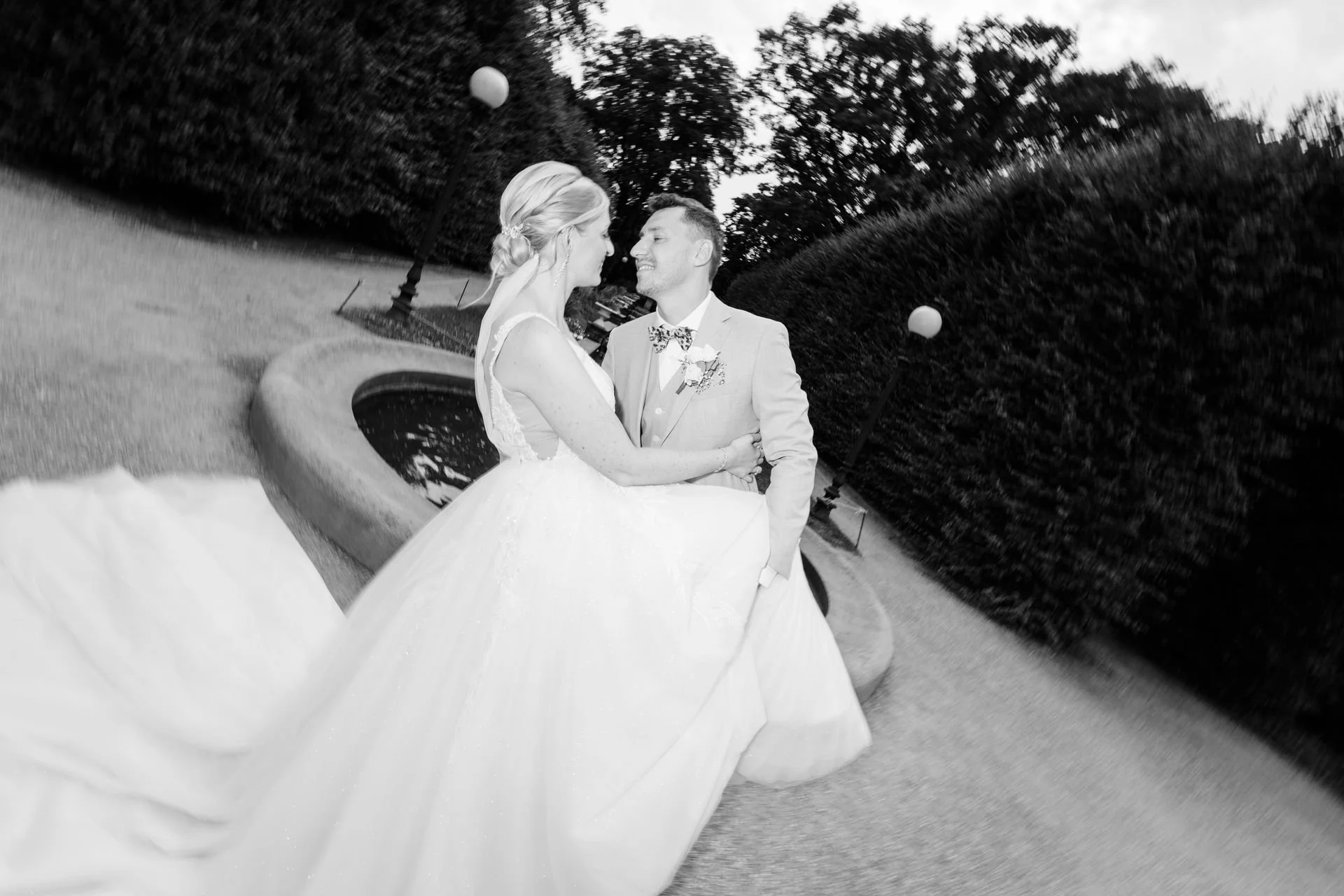 Braut und Bräutigam im Park, der Braut trägt ein elegantes weißes Hochzeitskleid, der Bräutigam einen Anzug mit Fliege, sie lächeln sich an, die Szene ist in Schwarz-Weiß aufgenommen.