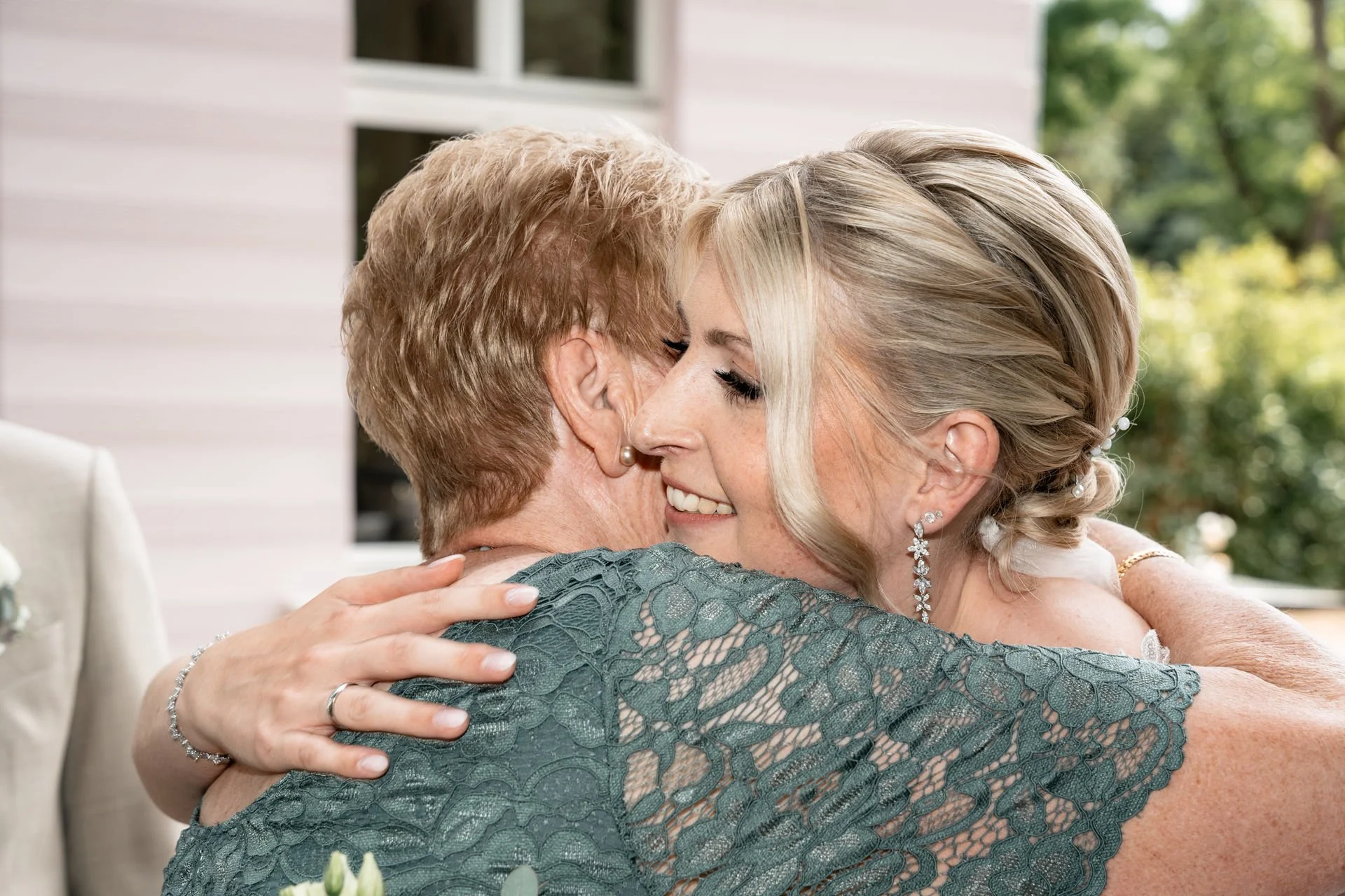 Zwei Frauen umarmen sich liebevoll, vermutlich bei einer Feier, im Hintergrund sind Bäume und ein helles Gebäude sichtbar.