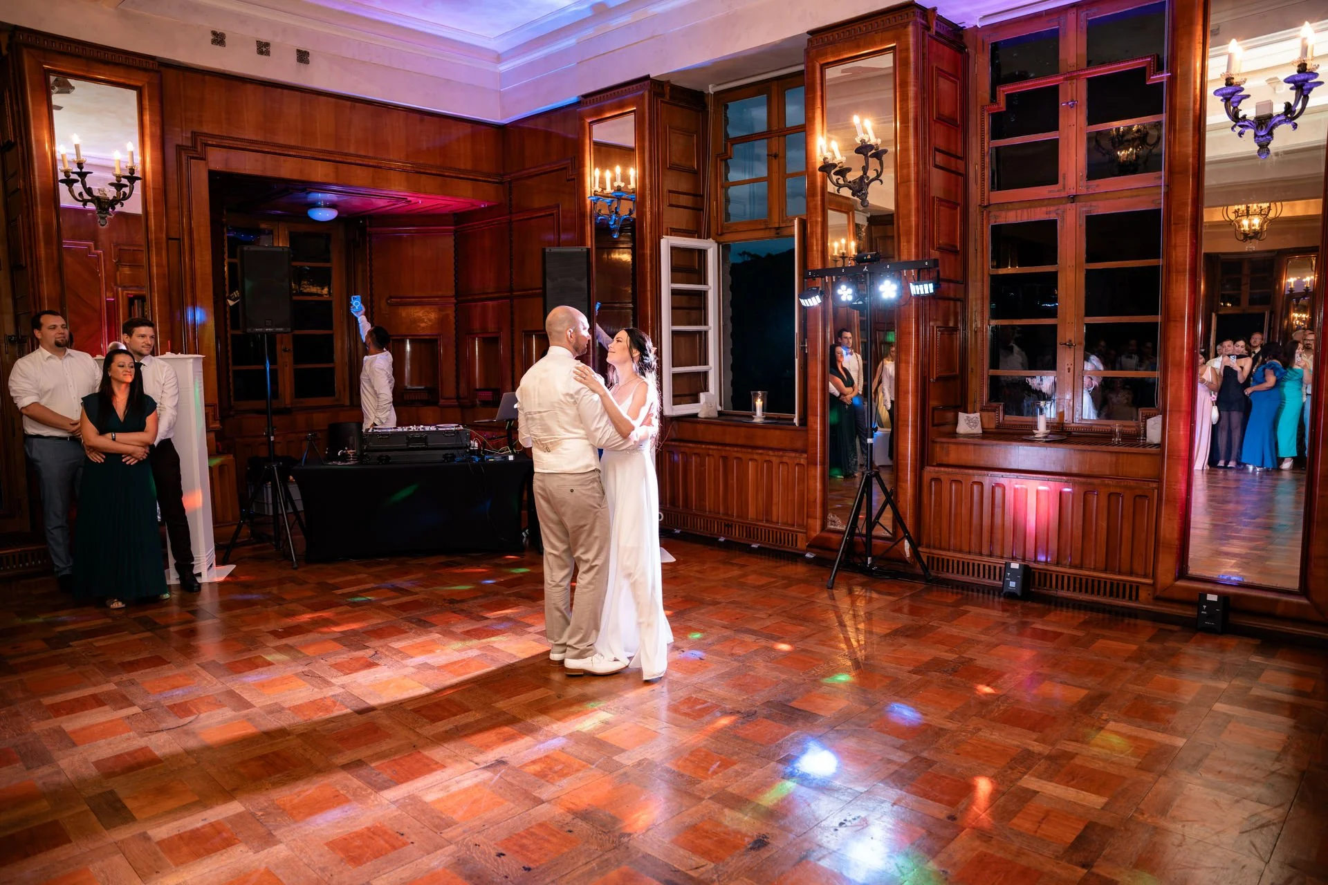 Ein Hochzeitsdoppel tanzt im festlich dekorierten Raum mit dunklen Holzvertäfelungen, großen Spiegeln und Kronleuchtern, während Gäste zuschauen.
