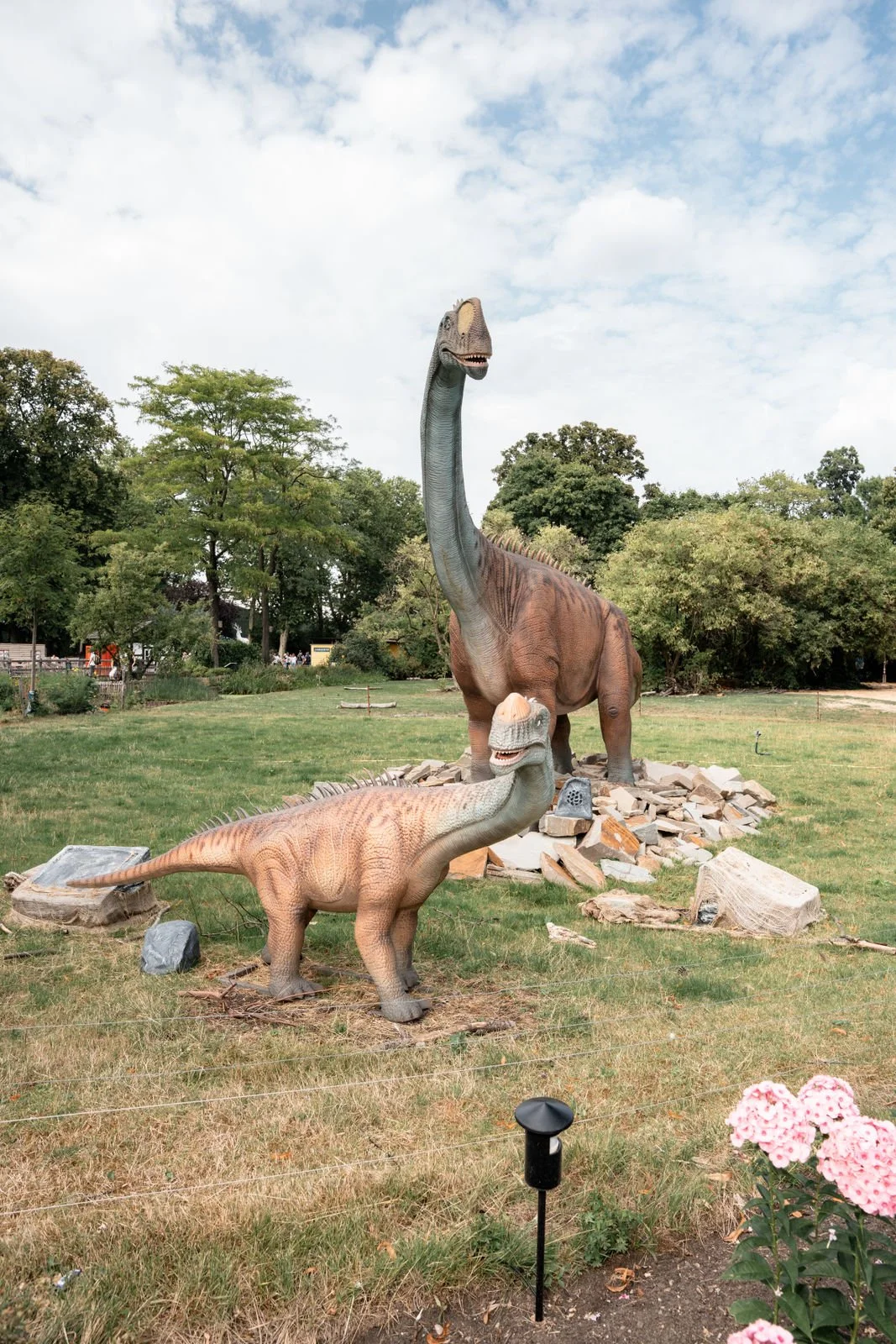 Dino-Dekoration im Park mit einem großen braunen Sauropoden und einem kleineren, hellbraunen Dinosaurier, umgeben von Bäumen, Steinen und Blumen, bei sonnigem Himmel.