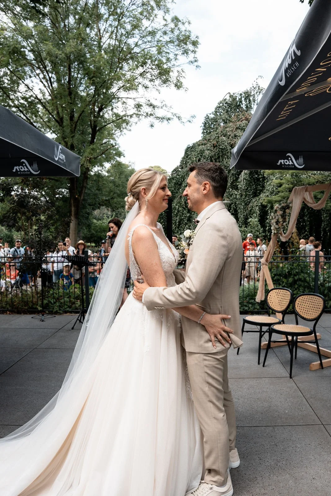 Ein heiratendes Paar beim Hochzeitswalzer im Freien, umgeben von Zuschauern und Bäumen.