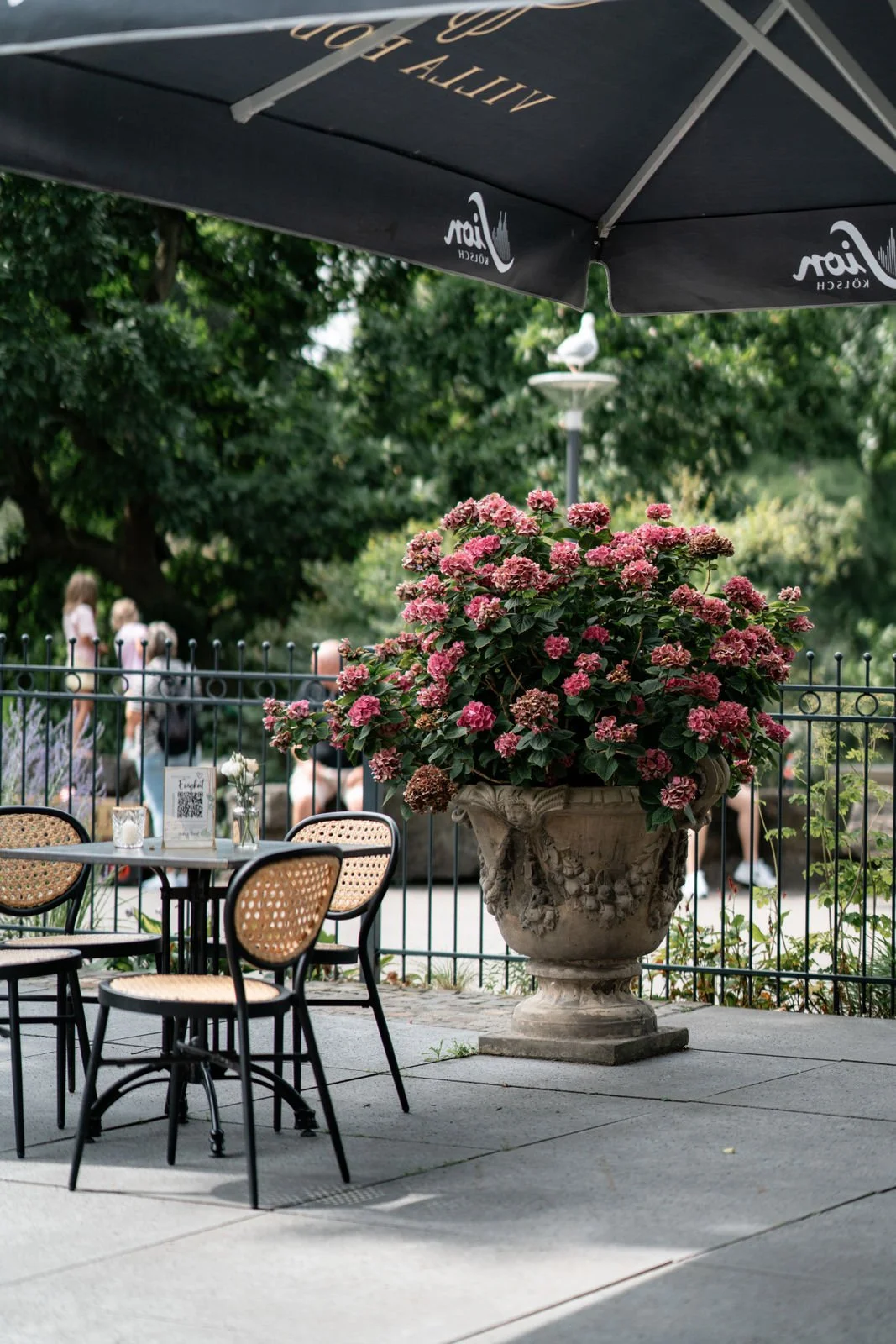 Ein Tische mit Stühlen im Außenbereich eines Cafés, eine große Blumenvase mit pinken Blumen, ein brauner Sonnenschirm, einige Leute im Hintergrund, und ein Vogel auf einer Laterne im Park.