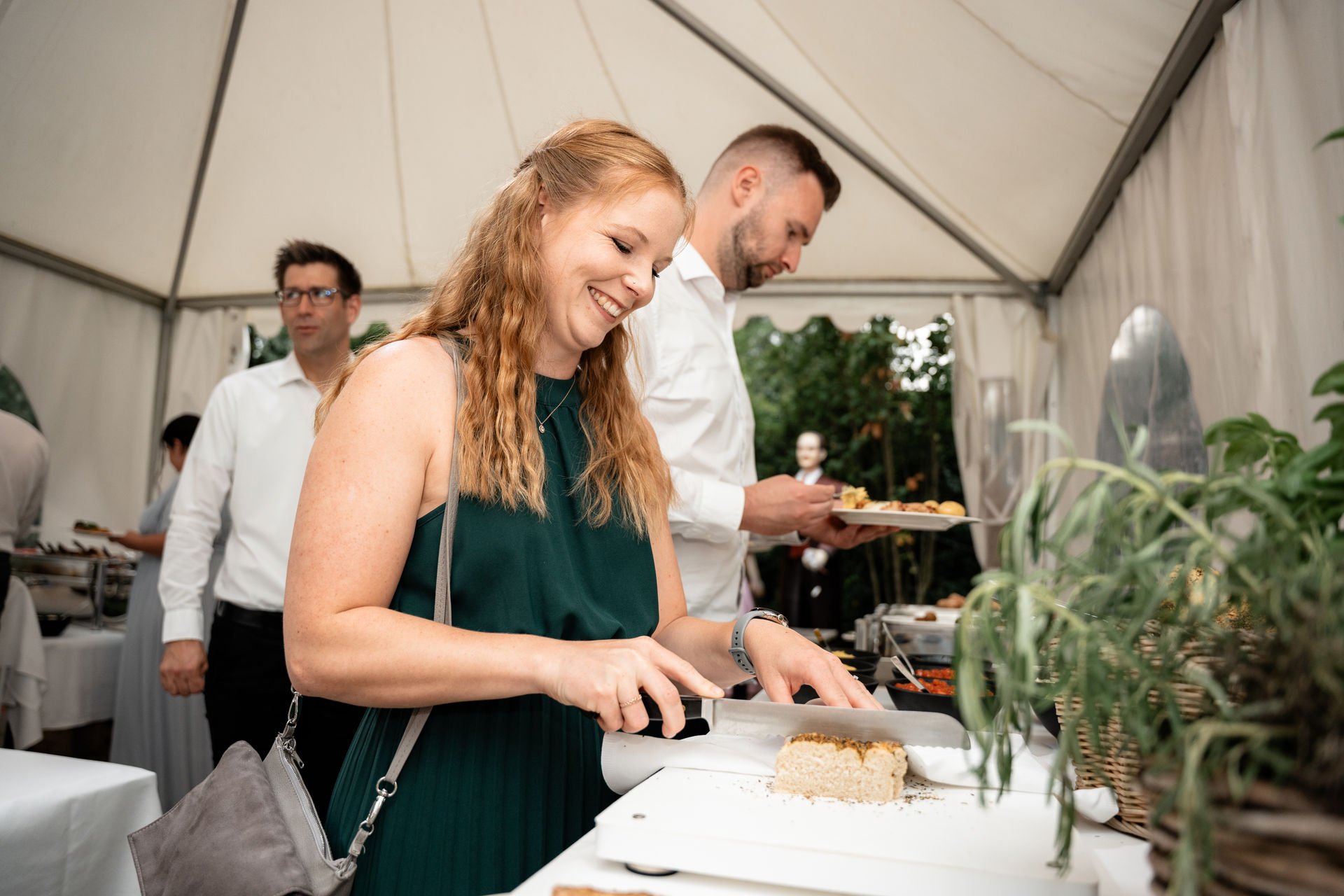 Frau schneidet Kuchen an einem Buffet auf einer Feier im Zelt, im Hintergrund weitere Gäste an einem Buffet.