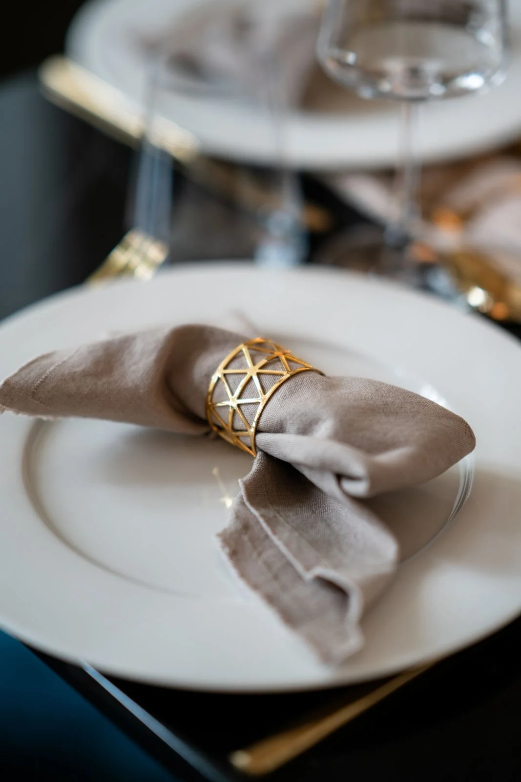 Eleganter gedeckter Tisch mit weißem Teller, beige Stoffserviette in Goldring, Weingläser im Hintergrund.
