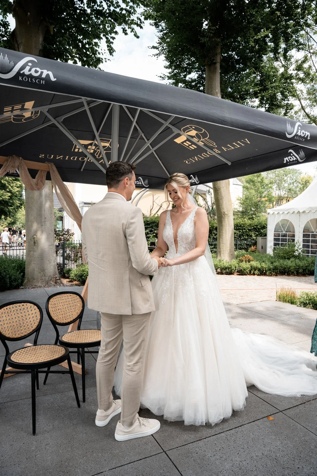 Ein Brautpaar bei ihrer Heiratszeremonie im Freien, unter einem großen schwarzen Sonnenschirm mit der Aufschrift 'VITAL PADDINUS' und 'Fion Kölsch'. Die Braut trägt ein elegantes, weißes Brautkleid mit tiefem V-Ausschnitt, und der Bräutigam trägt ein