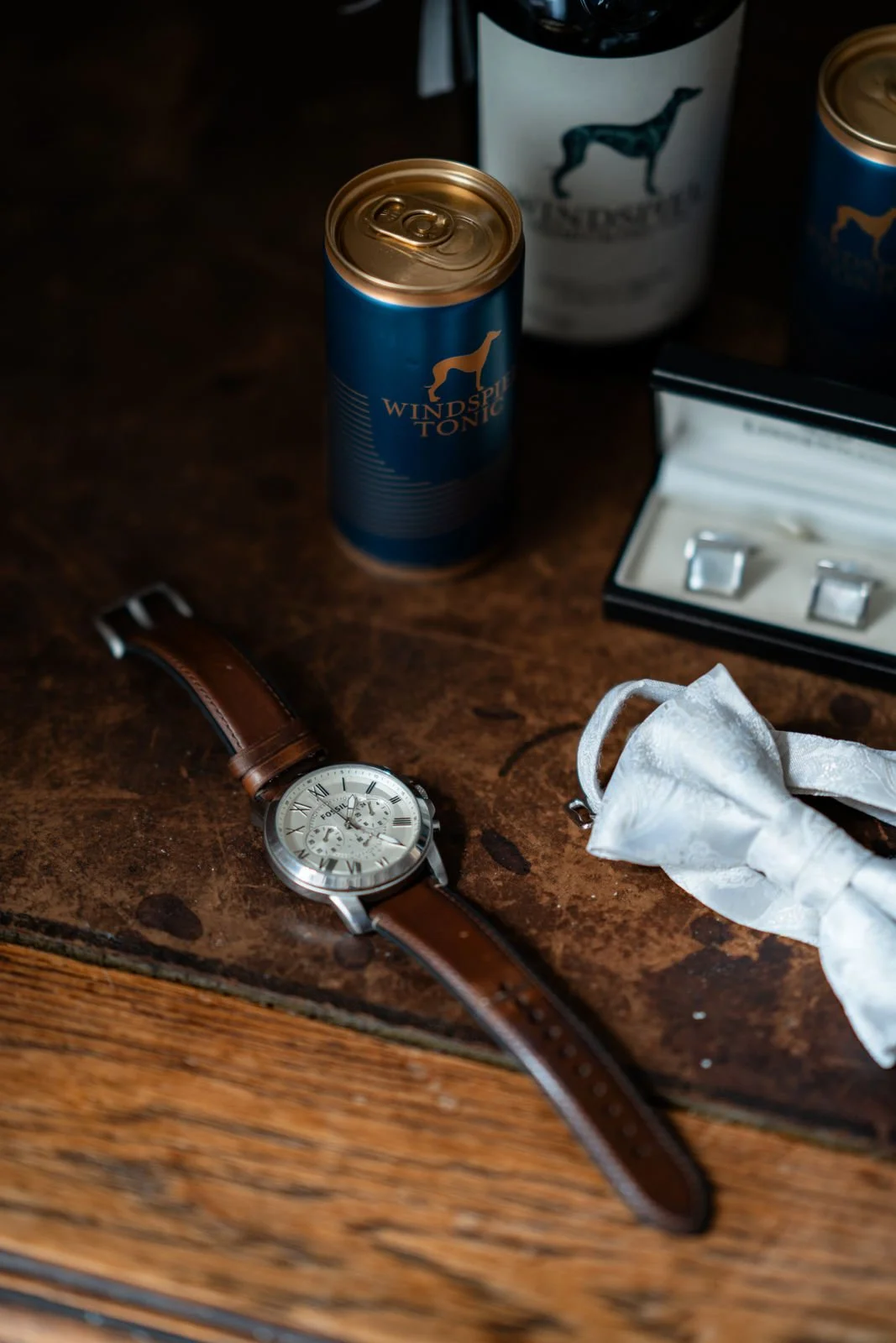 Eine Armbanduhr mit braunem Lederarmband liegt auf einem Holz Tisch neben einer Windspiel-Tonico-Dose, einer Flasche mit Giraffen-Design, einem schwarzen Karton mit Schmuckringen und einer aufgerollten Schutzmaske.