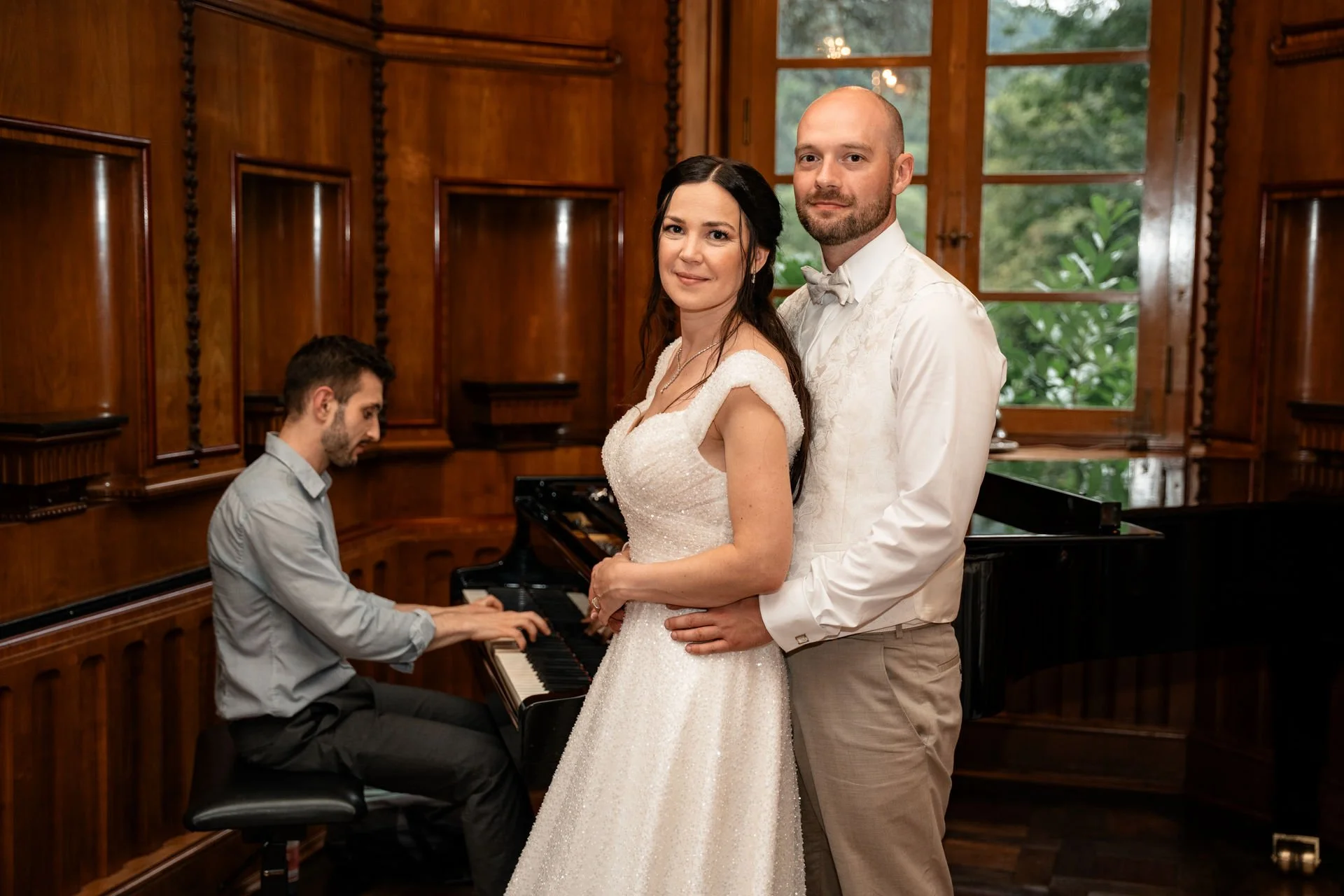Ein Brautpaar steht vor einem Klavier, während ein Pianist spielt, in einem Holzsaal mit großen Fenstern im Hintergrund.