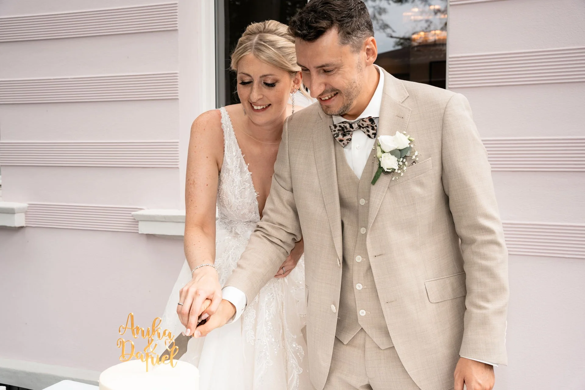 Ein Brautpaar schneidet gemeinsam die Hochzeitstorte an. Die Braut trägt ein weißes Hochzeitskleid, der Bräutigam ein hellbeiges Jackett mit Fliege. Beide lächeln glücklich.