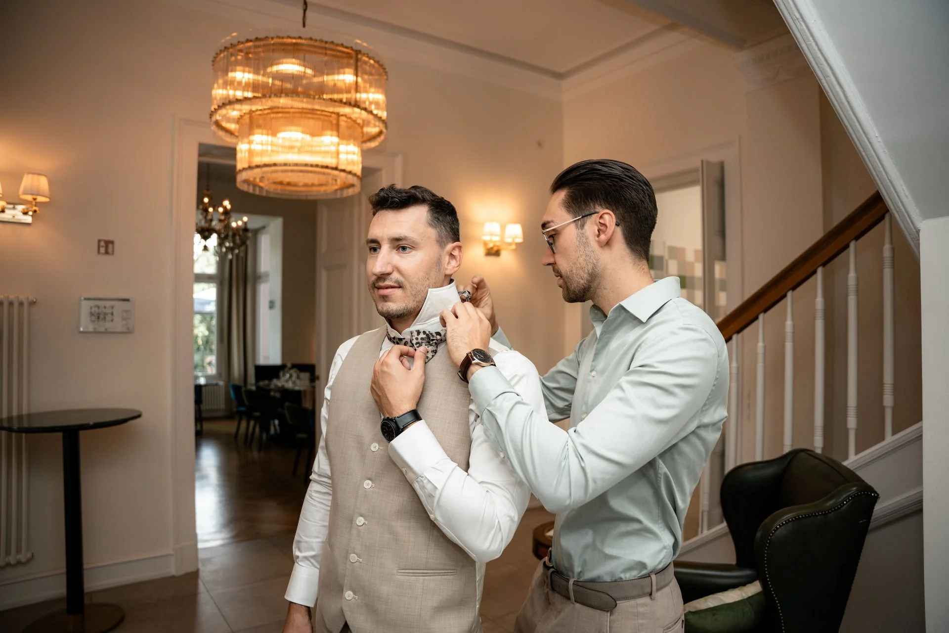 Zwei Männer bereiten einen Mann für eine formelle Veranstaltung vor, indem sie ihm ein Halstuch umlegen in einem eleganten Raum mit Kronleuchtern und stimmungsvoller Beleuchtung.