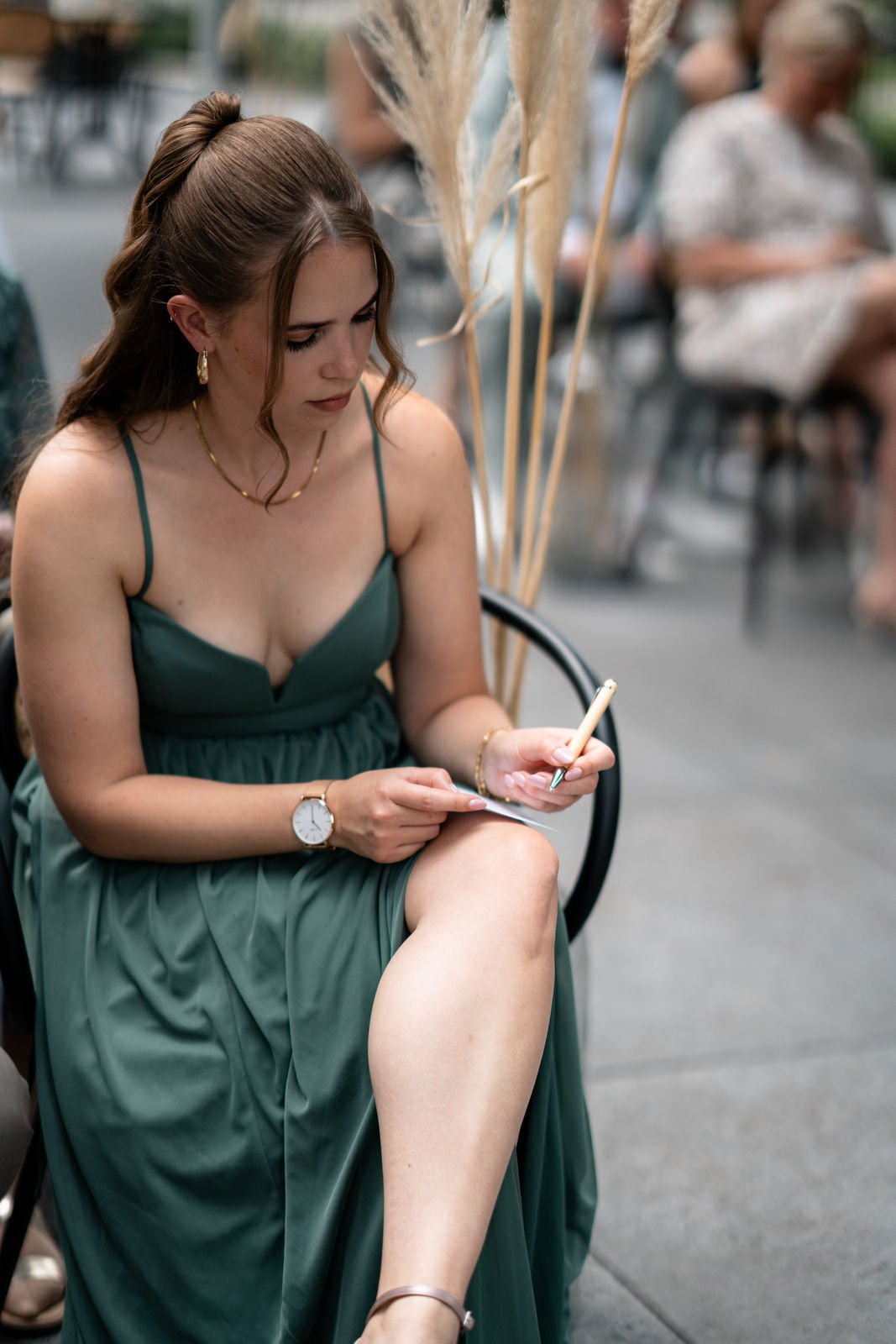Eine junge Frau in einem grünen Kleid sitzt auf einem Stuhl im Freien, liest oder schreibt in einem kleinen Notizbuch, trägert Uhren und Schmuck, umgeben von anderen Personen, die im Hintergrund sitzen.