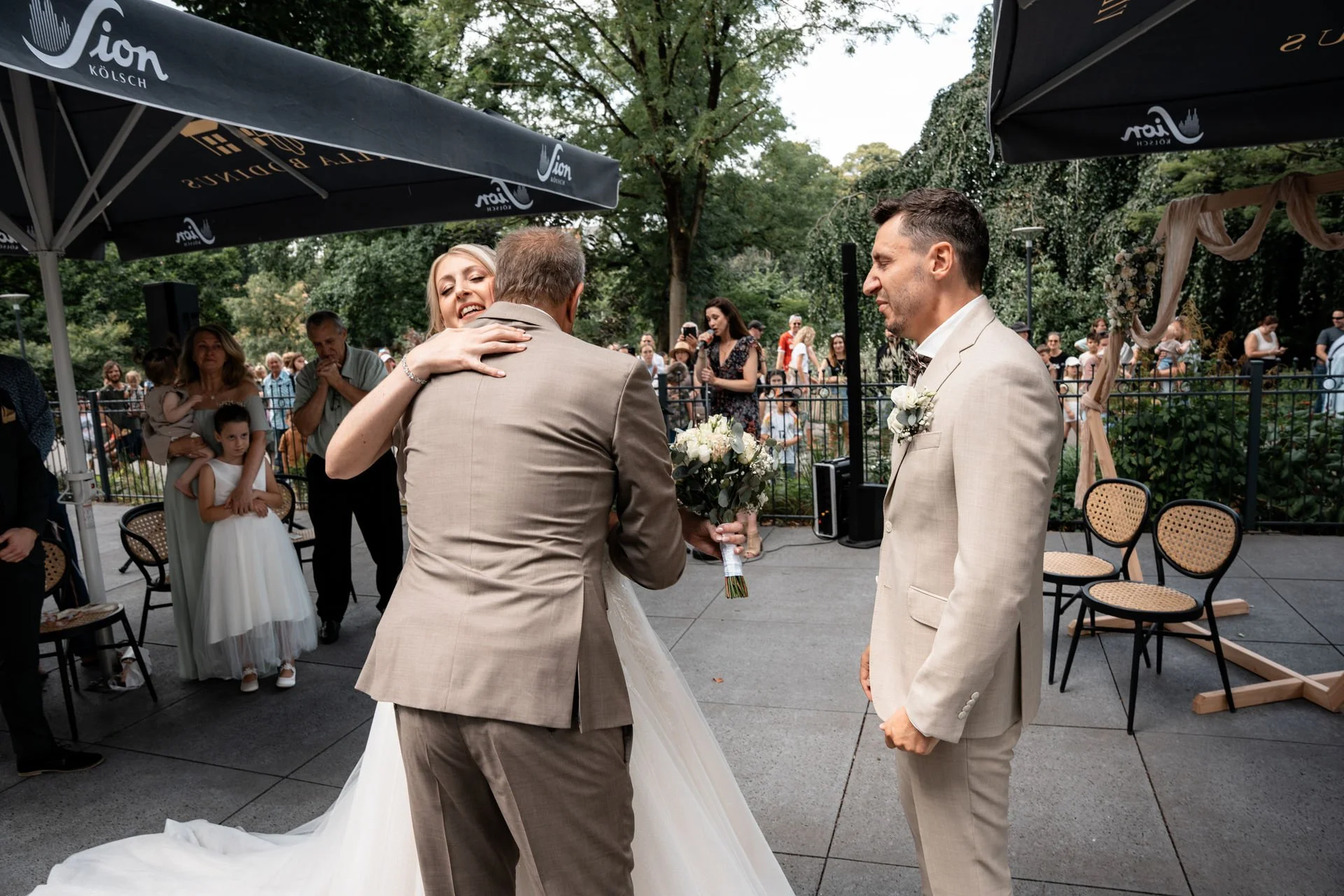 Hochzeit im Freien, Braut küsst den Bräutigam, der einen Blumenstrauß hält, umgeben von Gästen, unter einem schwarzen Sonnenschirm.