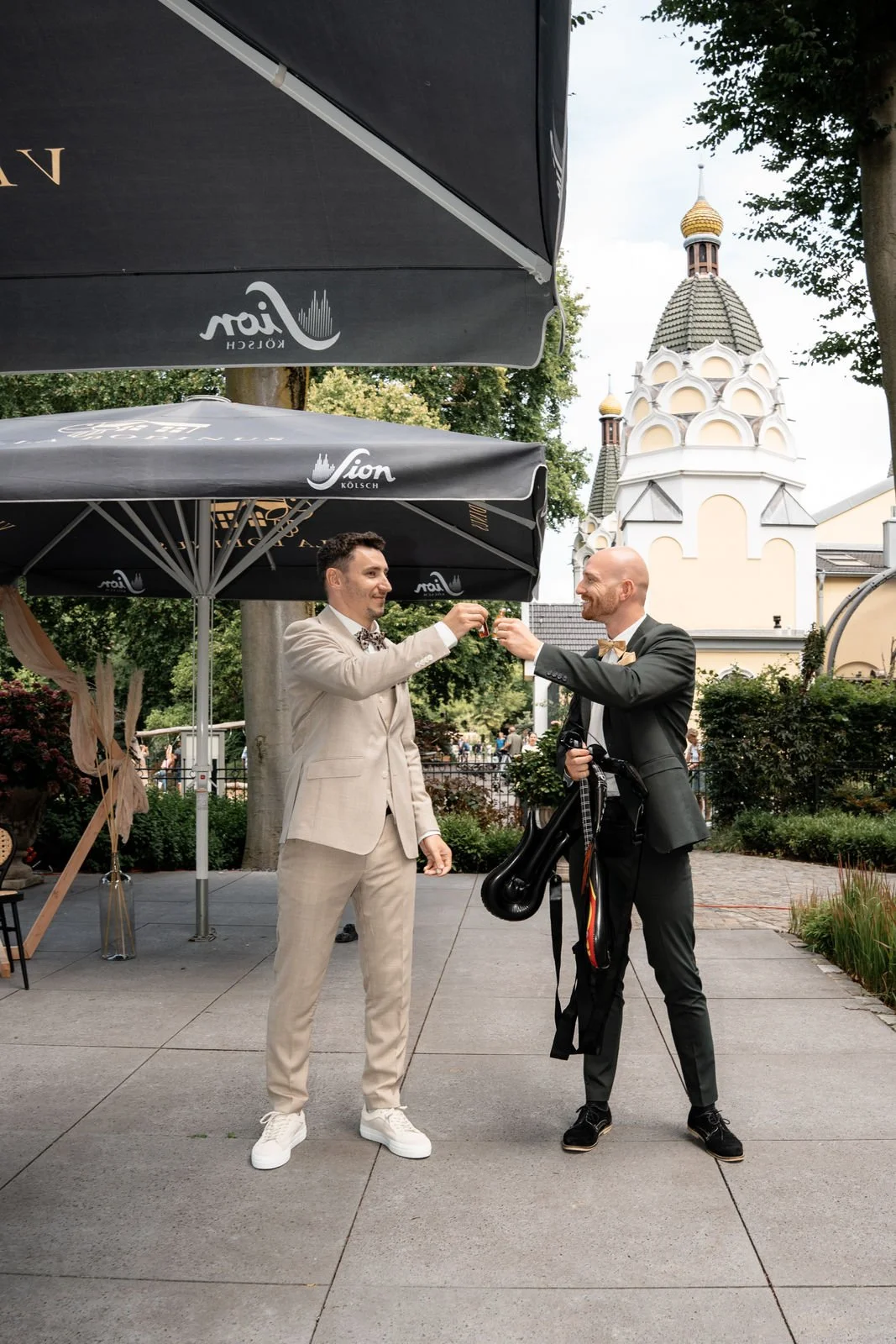 Zwei Männer in Anzügen stoßen sich mit Gläsern an, einer hält eine Traukarte, der andere trägt eine Golf-Tasche. Ein Gebäude mit einer europäischen Kirche im Hintergrund ist sichtbar.
